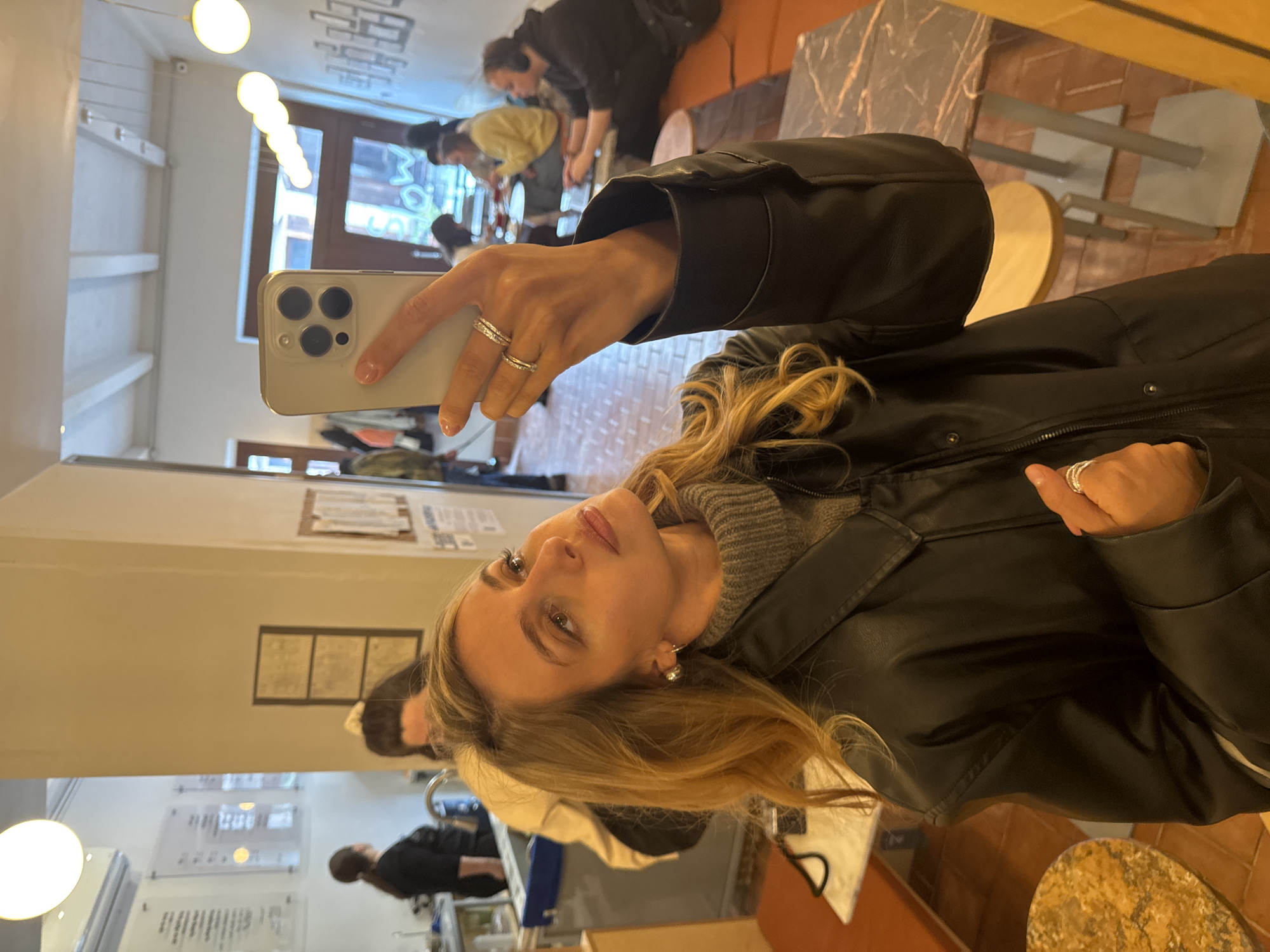 The image is a mirror selfie, rotated 90 degrees counter-clockwise, featuring a woman in an indoor setting, likely a cafe or restaurant in Tbilisi, Georgia.
The primary subject is a woman with long, wavy blonde hair, light-colored eyes, and full lips, looking directly at her reflection with a neutral expression. She is wearing a black leather-like jacket over a grey ribbed turtleneck or sweater. Her left hand, holding a light-colored iPhone with three camera lenses, prominently displays two rings on her ring finger. Her right hand is also visible, adorned with multiple rings.
The background, seen in the mirror's reflection, reveals a bustling but out-of-focus interior. Several other patrons are present; some are seated at tables, while one individual in a yellow top is bending over. The setting includes tables with what appear to be marble-patterned tops and chairs, suggesting a public eating or gathering space. The floor is tiled with warm-toned materials. The ceiling features several spherical light fixtures and a rectangular light panel, providing warm illumination. White shelving and indistinct framed documents or signs are visible on the walls. Through a window, some blurry, unreadable signs are reflected from outside. The scene depicts a casual gathering in a well-lit establishment.