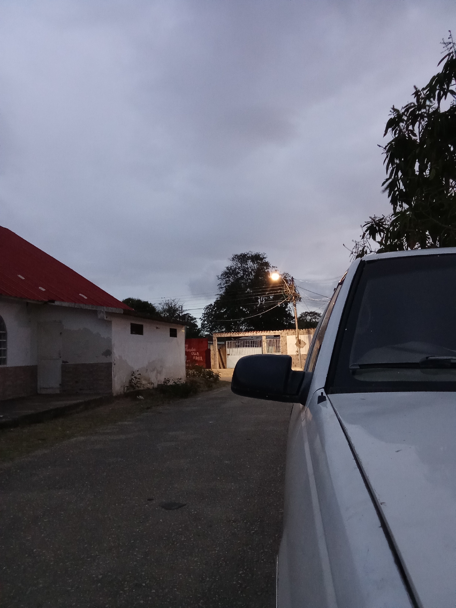 The image depicts a street scene in Juan Griego, Venezuela, taken at dusk or dawn. A white building with a red roof stands to the left of the frame. The street is paved and leads toward a lit street lamp in the distance. A silver or white car is parked on the right side of the road, taking up a significant portion of the right side of the frame. A tree is visible behind the car. The sky is overcast. In the distance, the architecture of buildings suggests a residential area. No people are visible.