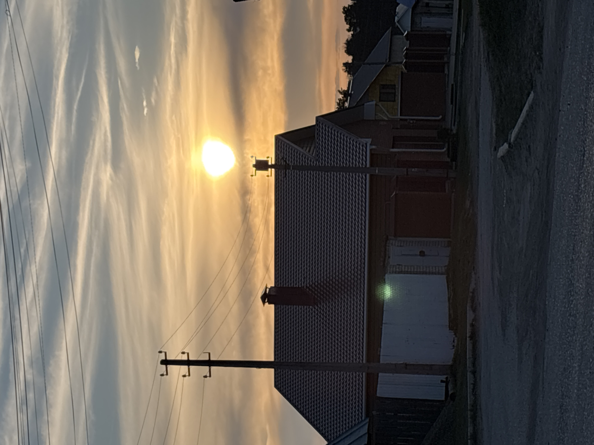 The image shows a sunset scene in what appears to be a residential area. A house with a grey-tiled roof and a chimney is prominently featured in the middle of the frame. A utility pole stands in front of the house, with several wires stretching across the sky. The sky is filled with clouds, colored in shades of orange and grey by the setting sun. The sun is a bright spot in the sky. Part of a road is visible on the right side of the image. Other houses and trees can be seen in the background. The location is indicated as Chaikovsky, Russia, based on the prompt.