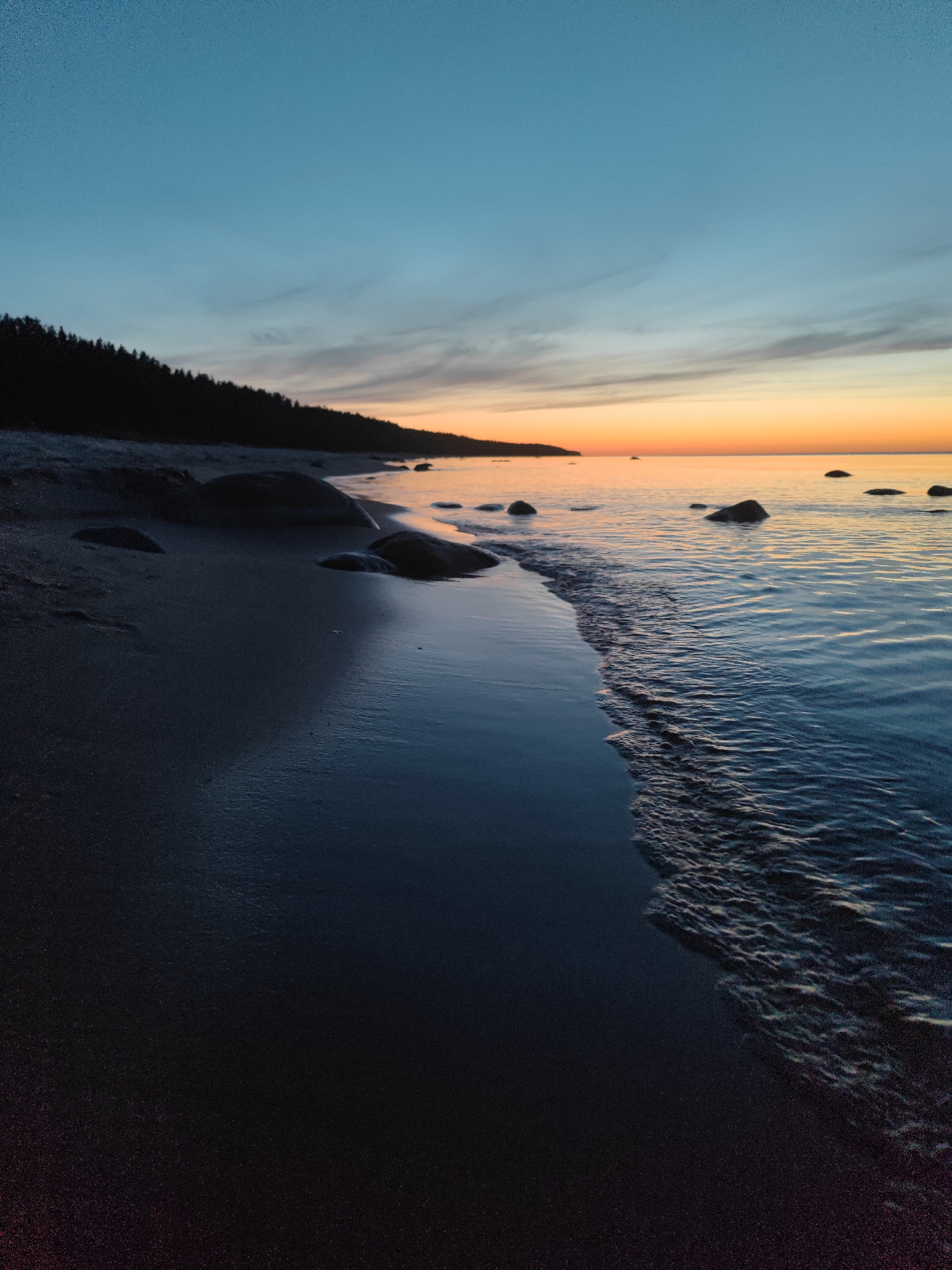 A tranquil beach scene unfolds at dusk, with a silhouette of a dense forest lining the horizon on the left. The sand is dark and speckled, with several large, dark rocks scattered along the shoreline. The ocean to the right is calm, reflecting the warm hues of the sunset that paint the sky in gradients of orange, yellow, and a soft blue. Small waves gently lap at the shore. The overall impression is one of peaceful solitude. The location is the city of Motornoe, Russia.