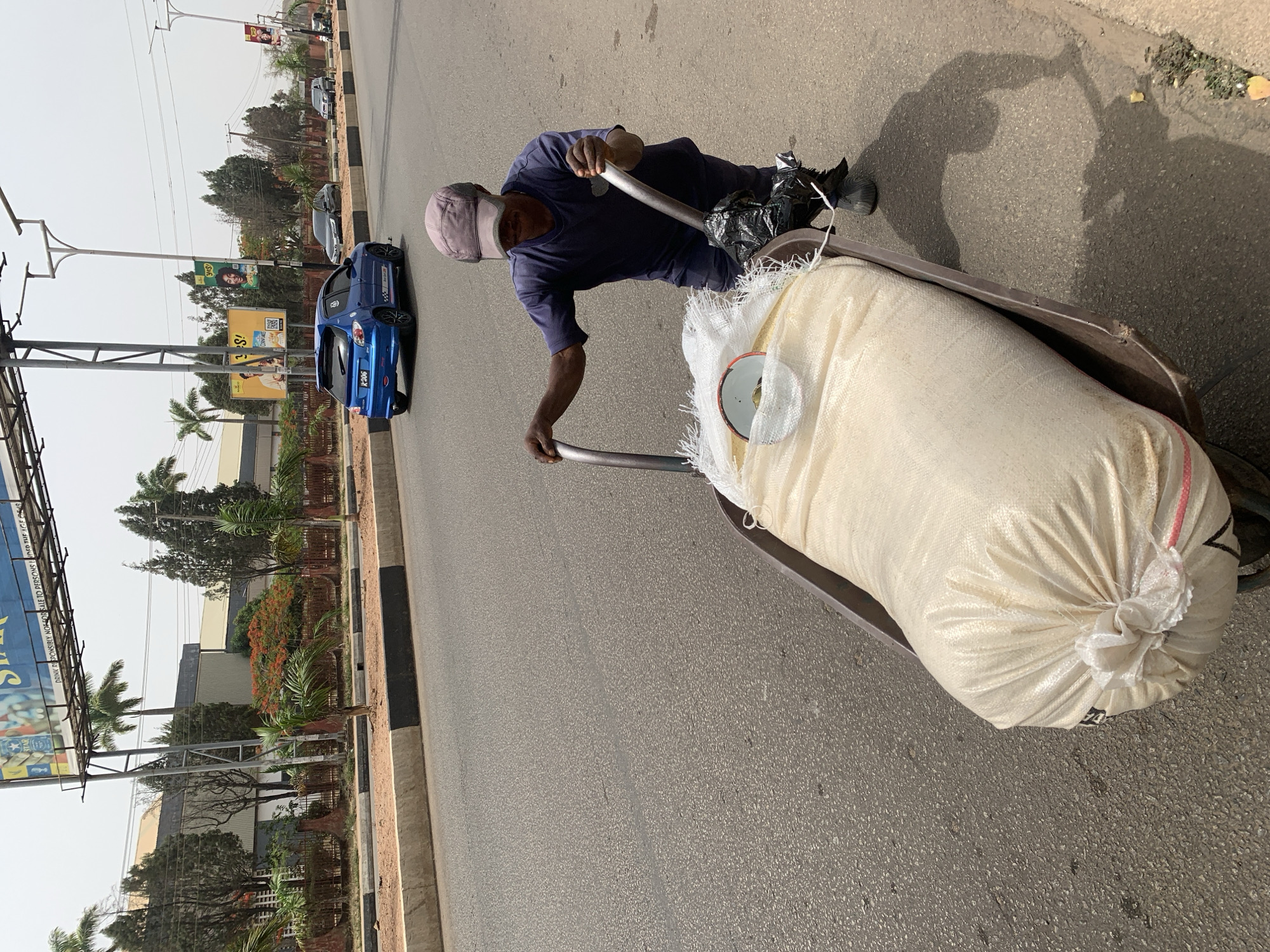 A man pulls a cart loaded with a large, white, tied sack down a street in Jos, Nigeria. The man is wearing a dark blue shirt, dark pants, a grey cap, and black boots. The cart is a metal container on wheels with a pull handle. A black plastic bag is also in the cart, near the handle. The street is paved and lined with a curb. Beyond the curb is a landscaped median with trees, flowering shrubs, and a billboard. A blue car is parked on the side of the road. The sky is overcast. The scene is in daylight.