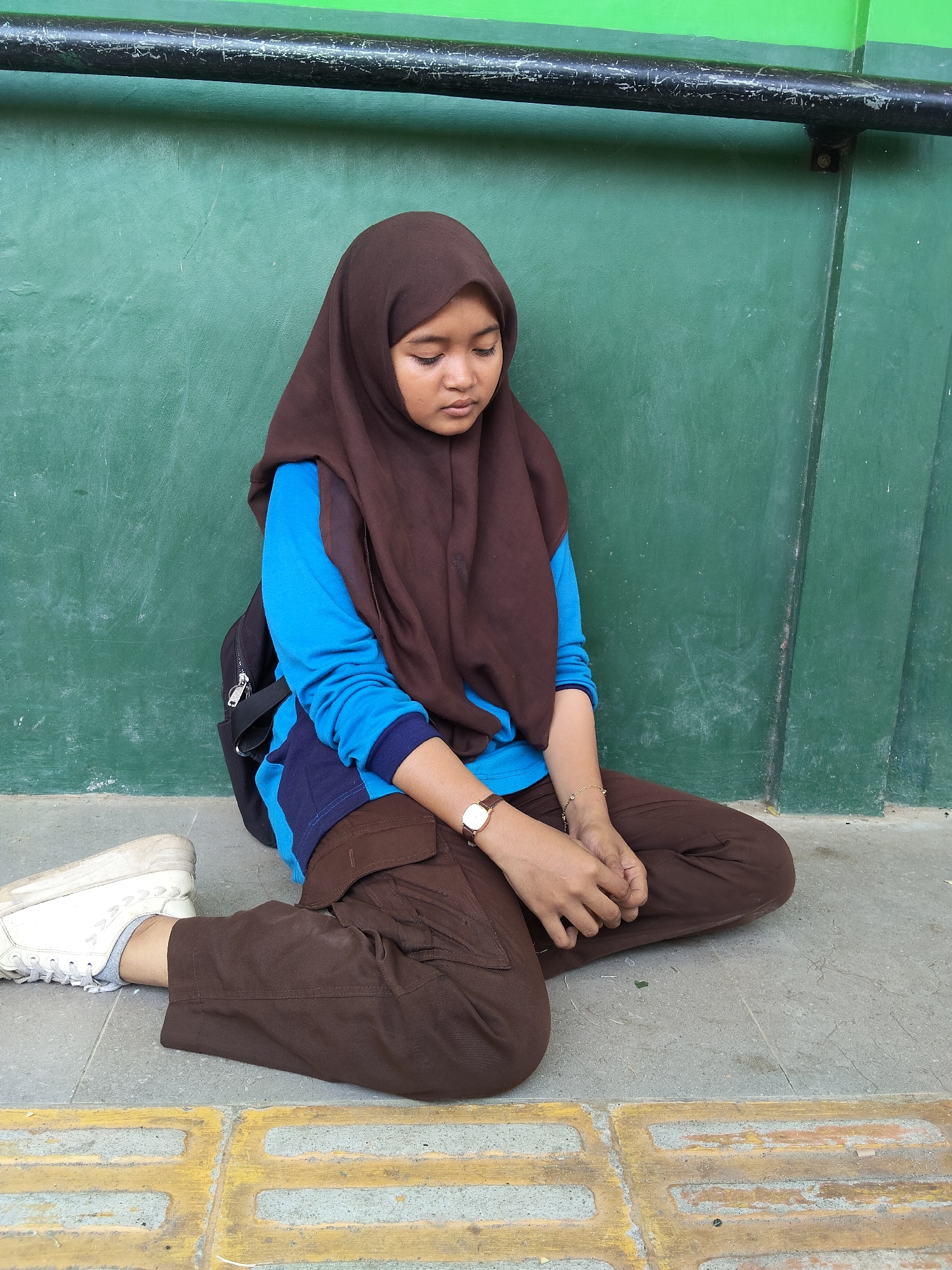 A young female, appearing to be of Indonesian descent, is seated on the ground in front of a green wall. She is wearing a brown hijab, a bright blue long-sleeved shirt, and brown cargo pants. A black backpack is visible behind her. She has a watch on her left wrist and a delicate bracelet on her right. Her hands are clasped in front of her, and she is looking downwards. The ground she is sitting on is paved, with yellow tactile paving blocks visible in the foreground. A metal pipe, painted black and showing signs of wear, runs horizontally along the top of the green wall. The scene is outdoors, likely in a public area within the city of Lori, Indonesia, given the context of the origin of the image.