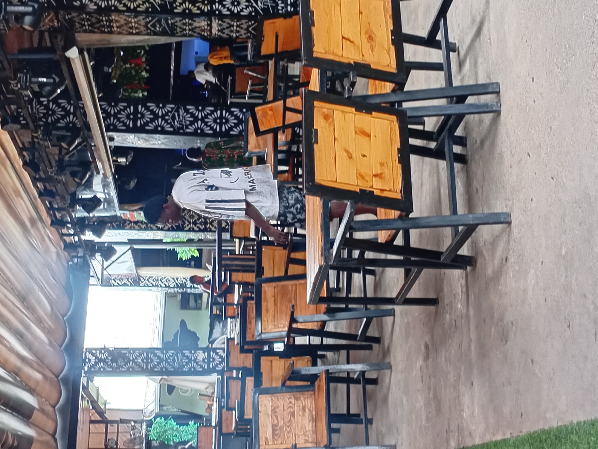 The image depicts an outdoor seating area, likely a restaurant or bar, with numerous empty tables and chairs. The furniture is made of wood with black metal frames, arranged on a concrete floor. In the background, there are decorative screens with floral cutouts and lighting fixtures. Several individuals are visible in the background, some appearing to be staff members. The overall atmosphere suggests a place that is either preparing to open or has recently closed for the day. The scene is set in Howayi, Nigeria, a location known for its vibrant nightlife and social establishments.