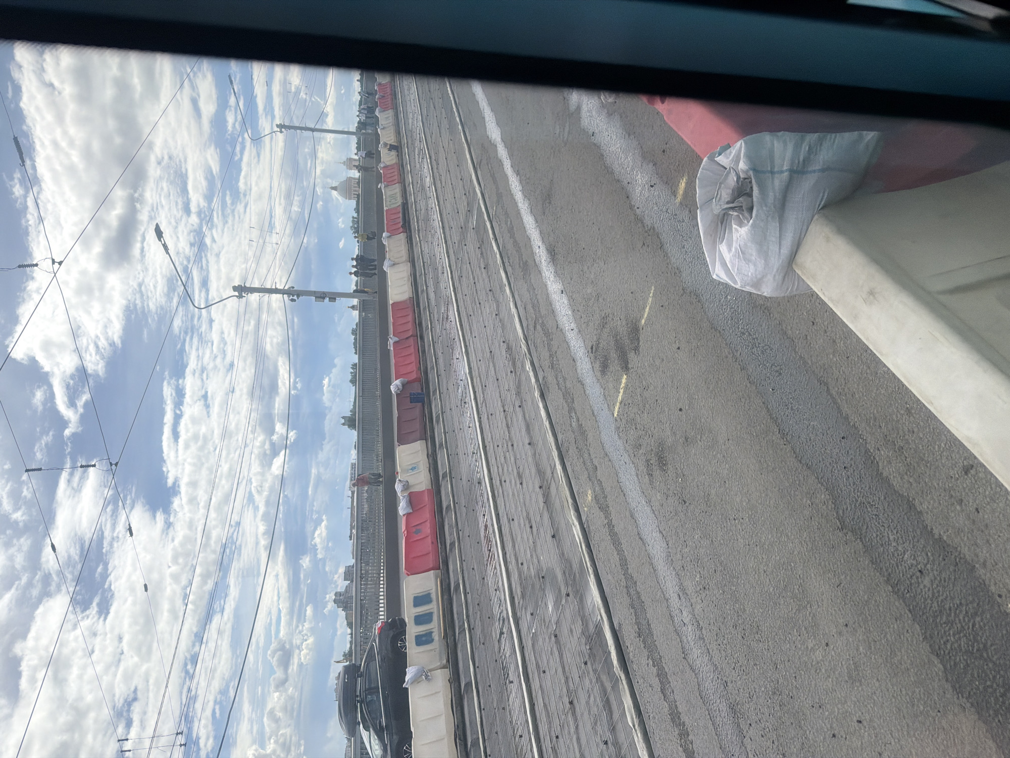 The media file captures a view from inside a vehicle, looking out at a construction site or roadwork. The scene is dominated by a road or pathway divided by lines of temporary barriers, some red and white, others white with blue markings. These barriers appear to be a type of traffic or construction barrier. In the foreground, a white fabric bag is placed on a raised concrete structure.
The background reveals a cloudy sky with bright patches, suggesting daytime. The sky is crisscrossed with overhead wires, possibly for electrical or communication lines, supported by utility poles. In the distance, a few buildings are visible, including one with a domed roof, hinting at a city or town setting. Several people can be seen on a walkway or bridge in the distance, and some cars are parked along the side of the road.
The overall impression is one of ongoing work or development in an urban environment under a dynamic sky. The presence of construction barriers and the parked vehicles suggests a functional area, possibly a street or a track for public transport. The weather appears to be fair with scattered clouds. No visible text is present in the media file. The image is oriented vertically, giving a sense of looking up or down along the scene.