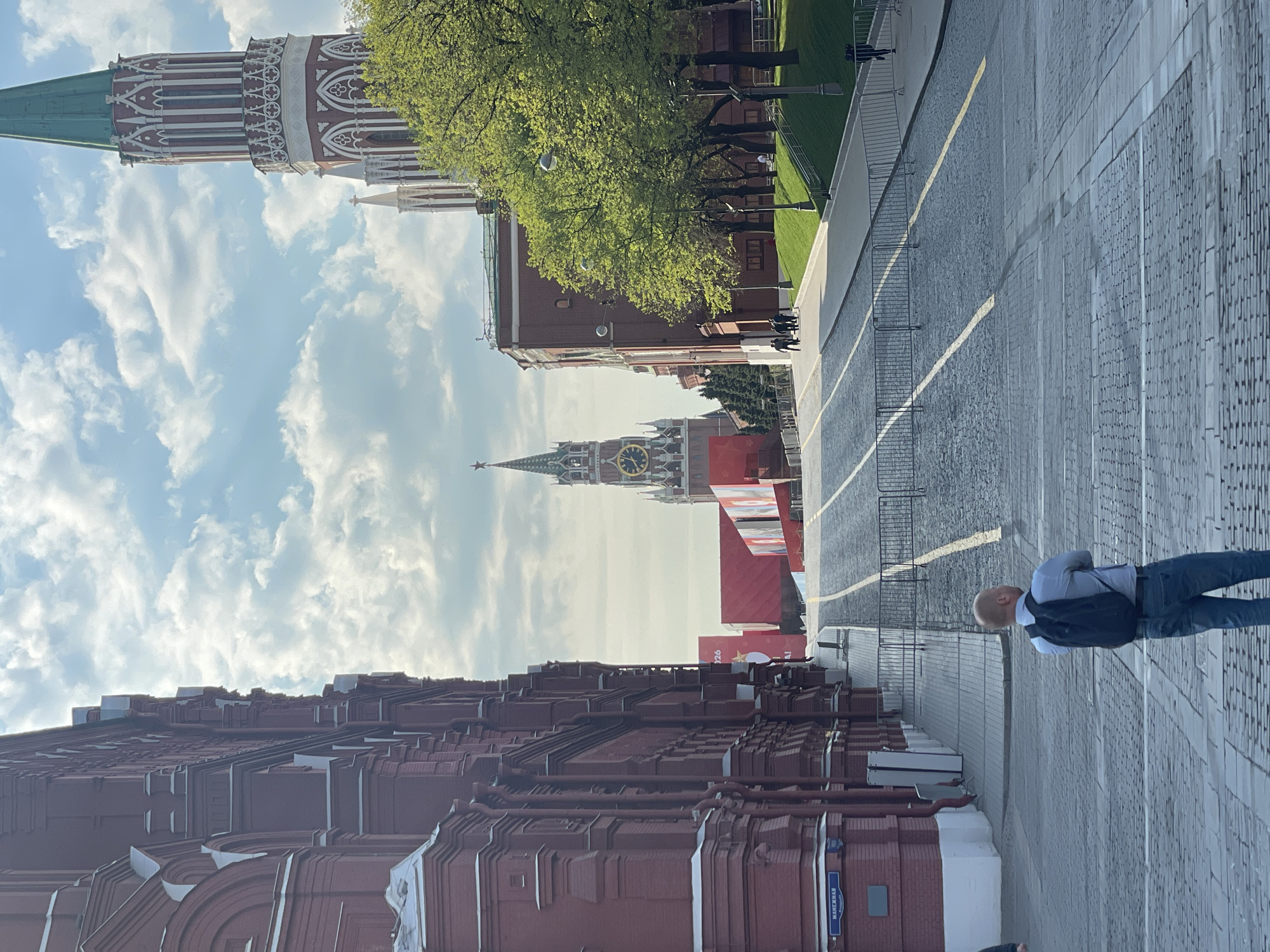 The image captures a partial view of the Moscow Kremlin complex in Russia, specifically focusing on the architectural details of the red brick walls and towers along with the Spasskaya Tower featuring its prominent clock face. Visible is a segment of the cobblestone pavement of Red Square, featuring a man in jeans and a light blue shirt standing on it. Metal barricades are also present. The sky is partly cloudy.