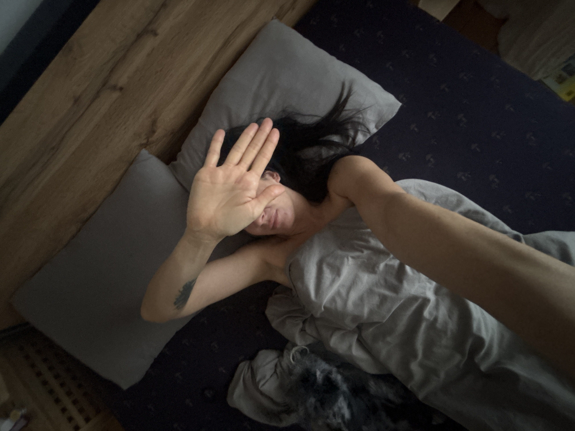 A woman is lying in a bed covered with a gray blanket, facing away from the headboard. She is partially obscured by the blanket, with a dark-haired head on a gray pillow and a hand raised to shield her face. A tattoo is visible on her left arm. The bed is made with gray pillows and a dark blue patterned bed sheet. The bed is situated near a wooden headboard and a wall. The image appears to be taken indoors. The location is not discernible as being Chaikovsky, Russia.