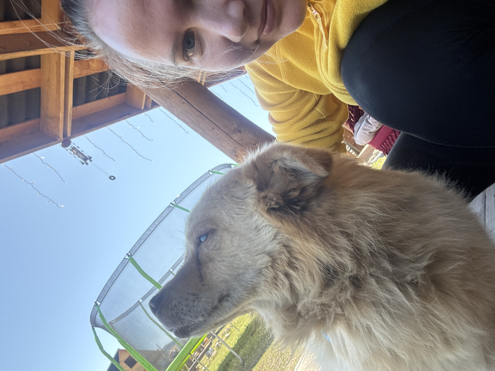 A woman wearing a yellow fleece jacket and black leggings is taking a selfie with a large, tan-colored dog. The dog is standing close to a green trampoline. The background includes the blue sky, a wooden structure overhead with strings of lights, and foliage. The image was taken in Tyulyuk, Russia.