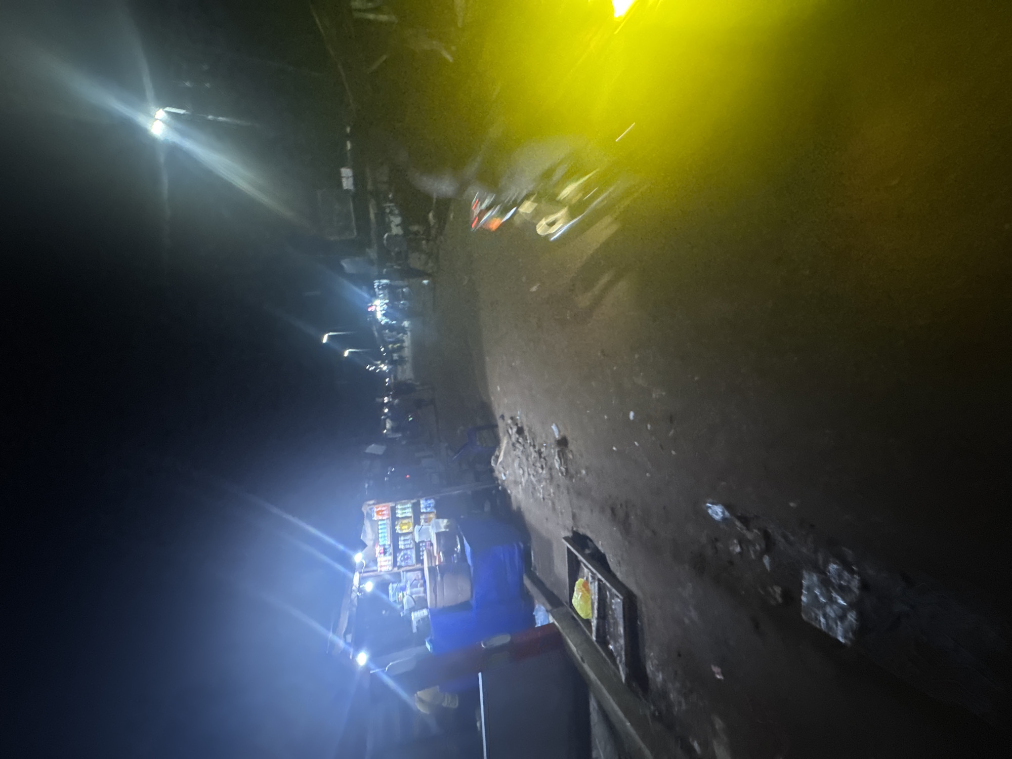 The image depicts a street scene in Ihiagwa, Nigeria, at night. Streetlights illuminate shops along the roadside. People and vehicles can be seen in the distance. A close-up view of what appears to be a roadside vendor stall is visible. A blue tarp covers some of the items on the stall.