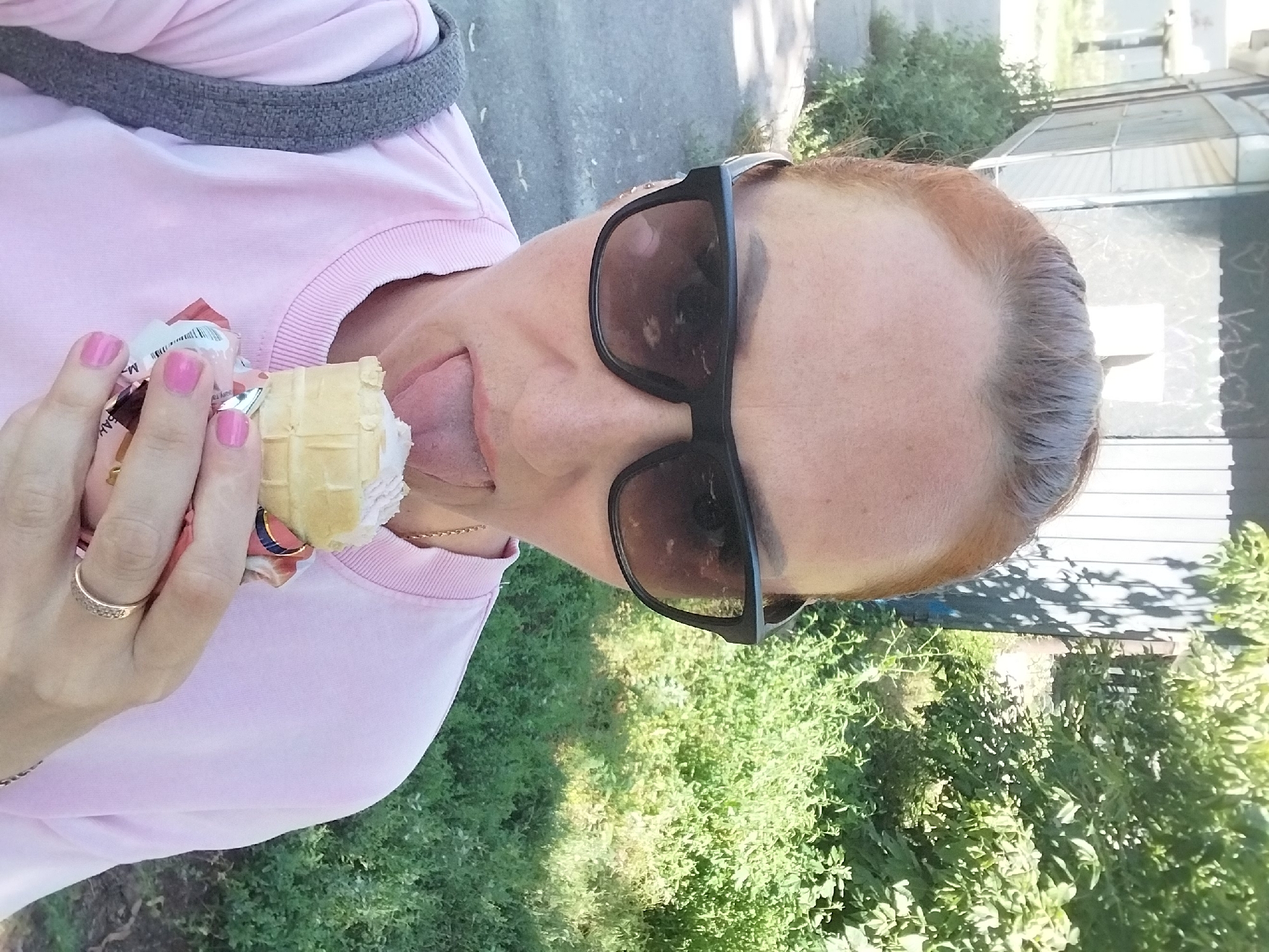 A woman, likely in her late 30s, with auburn hair pulled back, is pictured from the chest up, taking a selfie outdoors. She is wearing a light pink crewneck sweatshirt and large, dark sunglasses. Her tongue is extended, licking an ice cream cone in a waffle cone, held in her right hand. Her fingernails are painted a bright pink. A thin gold chain is visible around her neck. To her left, a portion of a gray strap from a bag is visible.
The background suggests an urban or suburban setting, with a building or structure on the left, partially obscured by greenery. The building appears to have a light-colored facade with dark, possibly spray-painted markings. Dense green foliage, including bushes and trees, dominates the right side of the frame. The ground in the foreground appears to be a paved surface, possibly a sidewalk.
The lighting indicates it is daytime, with bright sunlight casting shadows, suggesting an outdoor location. The overall scene is candid and informal.
The image was taken in the Dnipro city, Ukraine.