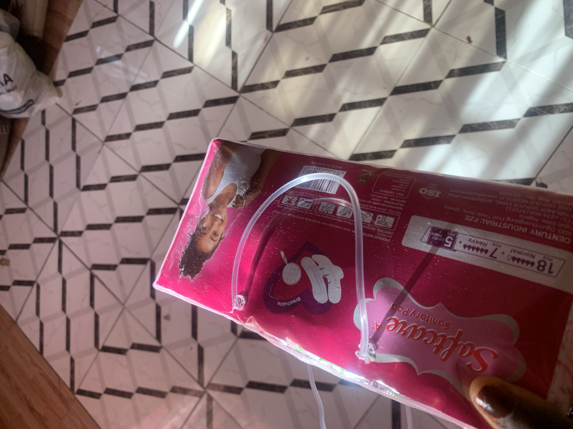 The image shows a package of "Softcare Sanitary Pad" positioned on a tiled floor with a geometric pattern of white and black squares. Sunlight streams in from the left, creating strong diagonal shadows across the floor and the product packaging. The packaging itself is primarily pink and features an image of a young woman with curly hair. Visible text on the packaging includes "Softcare Sanitary Pad," "MADE IN NIGERIA," "CENTURY INDUSTRIAL: FZE," "ISO 9001," and details about the contents and usage instructions such as "Normal," "Heavy," "panty liners," and "Breathable." The overall scene suggests a domestic setting, possibly a room in a home in Jauro Yinu, Nigeria, during daytime, given the strong sunlight and shadows. The product is a personal hygiene item, and the image's composition focuses on presenting the product itself.