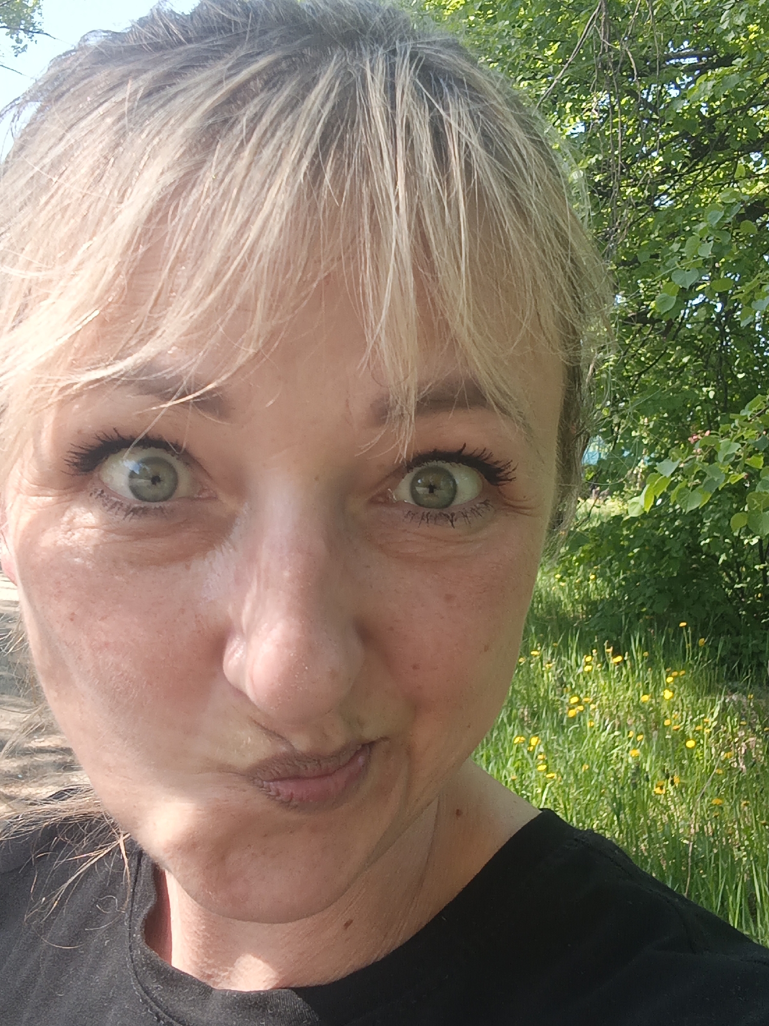 A close-up selfie photograph features a blonde woman with green eyes making a playful face, with her lips pursed and squinting. The woman is wearing a black t-shirt. The background shows green foliage, including trees and a field with yellow wildflowers. The location is the city of Chaikovsky, Russia.