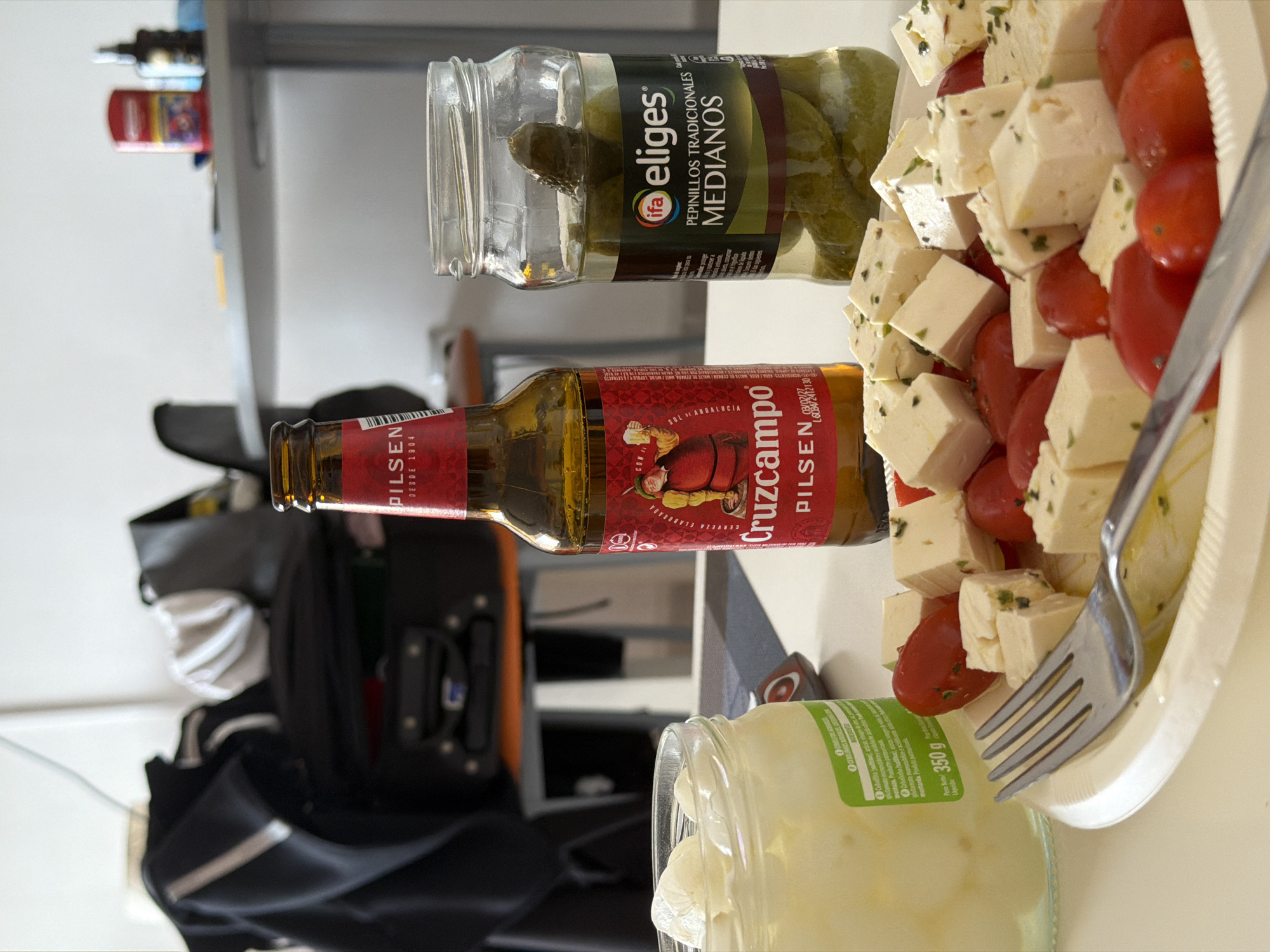 The image shows a table with various food items and drinks. From left to right, there's a black bag with compartments, a shelf with miscellaneous items, a bottle of Cruzcampo Pilsen beer lying on its side, a jar of pickles (eliges, pepinillos tradicionales medianos), and a platter of diced feta cheese and cherry tomatoes with a fork stuck into it. There is also a jar of 350 g of unknown contents. The objects are set on a light-colored surface, giving a snapshot of a possible meal or snack arrangement. Based on the products and branding visible, it is probably within Spain. Without additional geographic context or the presence of distinctive landmarks, it is impossible to pinpoint the exact location within Arroyo de la Miel-Benalmádena Costa, Spain.