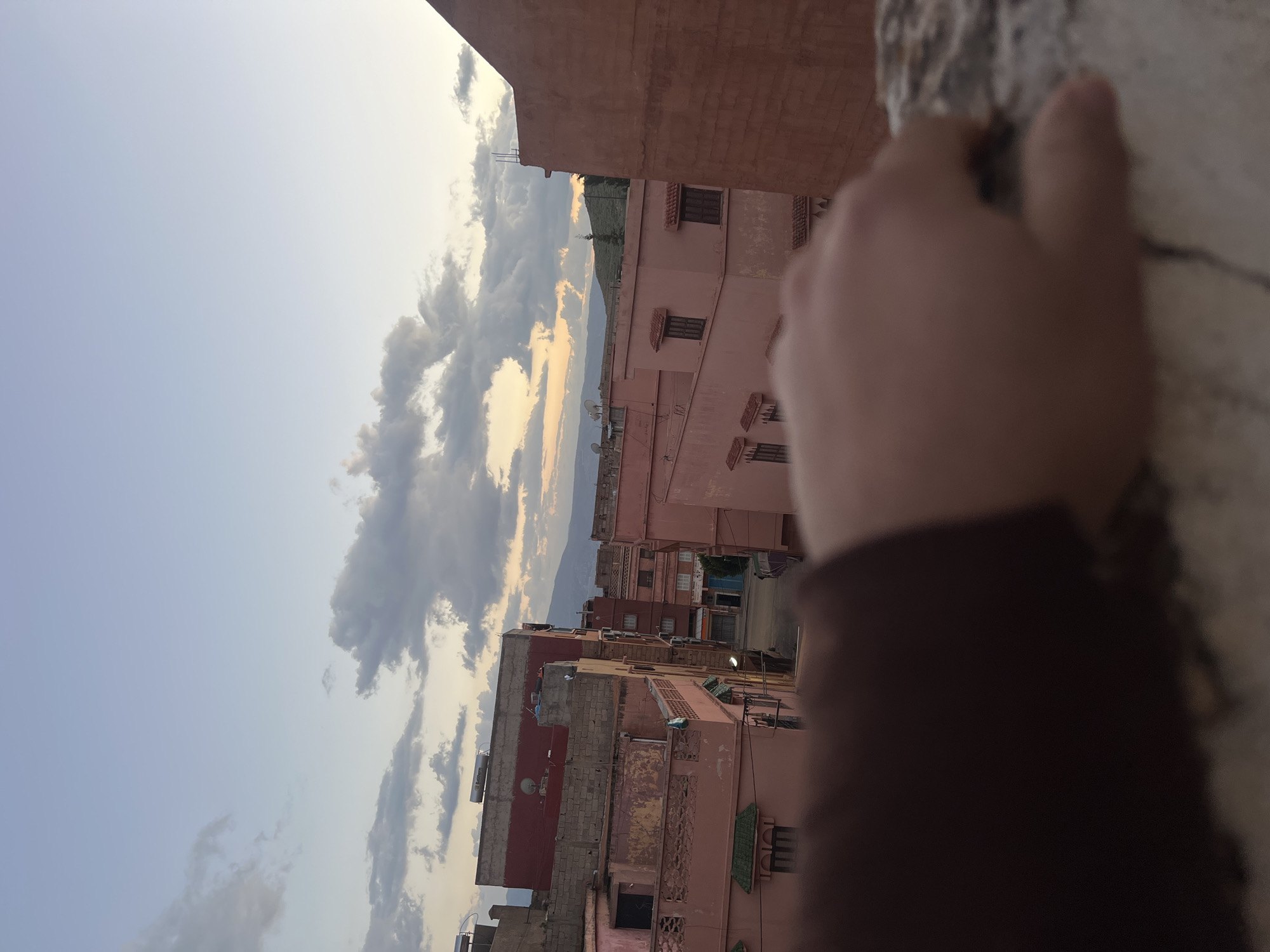 The image captures a sunset view in Ouaouizert, Morocco. Pink-hued buildings line a narrow street. Mountains are visible in the distance, framed by a cloudy sky tinged with the colors of the setting sun. A blurry hand and arm are positioned in the foreground on a wall or ledge.