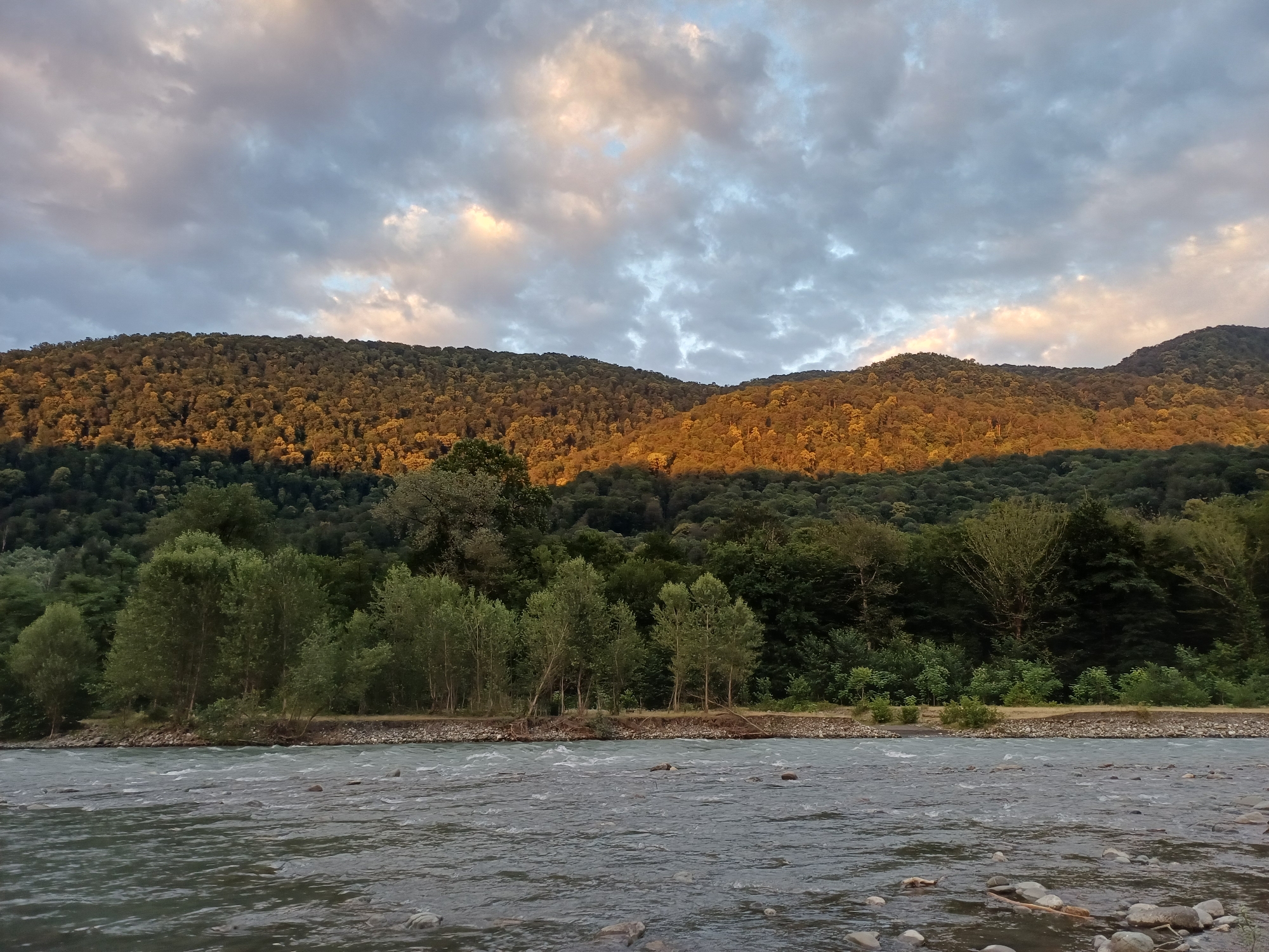 An outdoor, wide-angle, full shot captures a natural landscape scene by a flowing river under a cloudy sky. The river in the foreground has moderately turbulent water with visible rocks and debris. Beyond the river, a rocky bank transitions into a dense forest of varying shades of green trees. In the background, a large, forested mountain range stretches across the frame. The mountain slopes are partially illuminated by golden sunlight, contrasting with the shadowed areas. The sky above is filled with dramatic, gray and white clouds, with patches of warm, pinkish light suggesting a sunset or sunrise. The scene is devoid of human presence. The image was taken in Adlerskiy, Russia.
