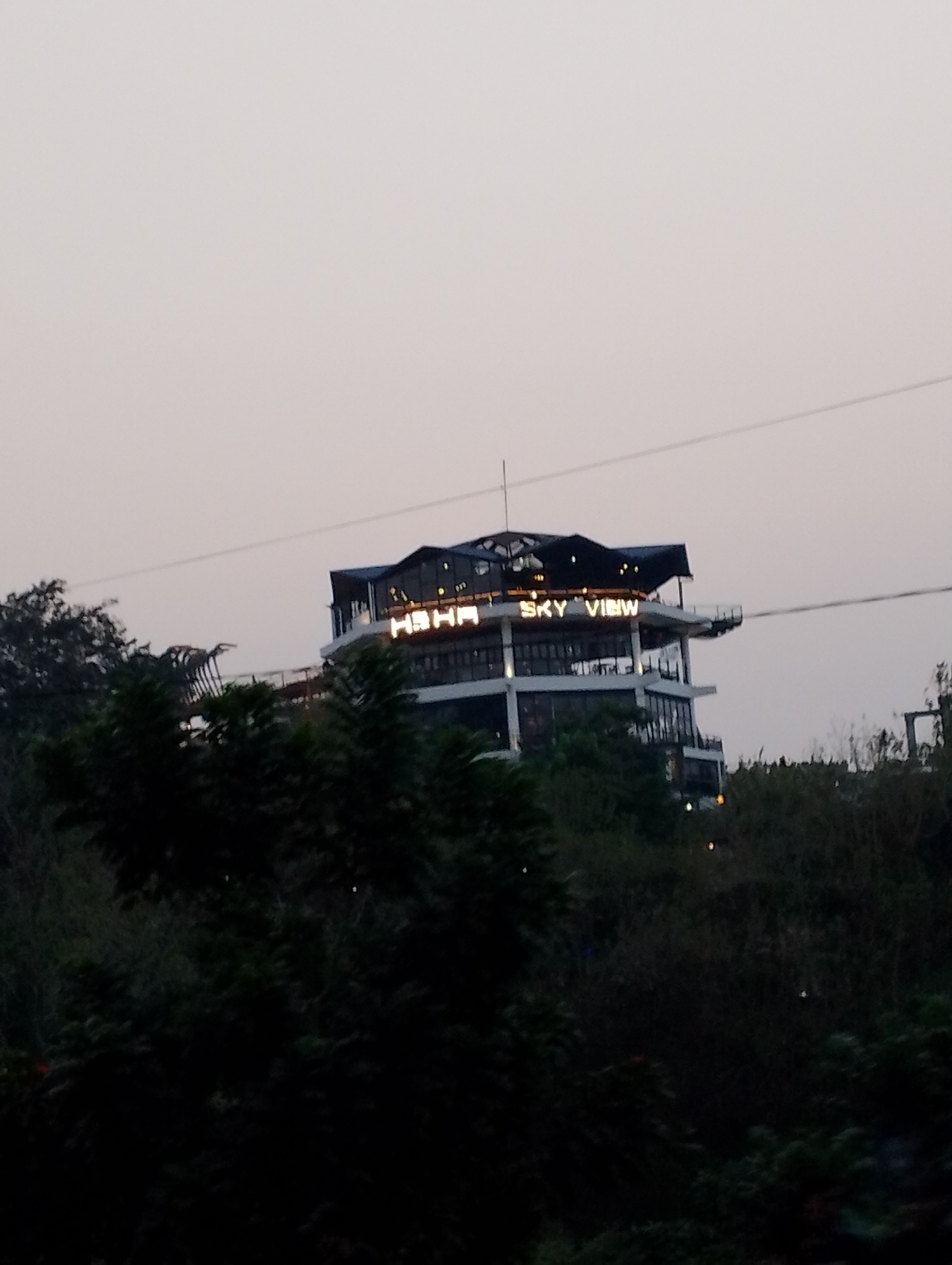 In Patuk, Indonesia, an image captures a multi-story building with illuminated signage that reads "HAHA SKY VIEW". The building appears to be a restaurant or observation deck, situated on a hillside surrounded by lush, dark green trees. The sky above is a pale, overcast grey, indicating either early morning or late evening light. A thin wire, possibly part of a zipline or antenna, extends horizontally from the building to the right. The building itself has a modern design with multiple levels, balconies, and large windows. The foreground is dominated by dense foliage, obscuring the base of the structure and the surrounding terrain.