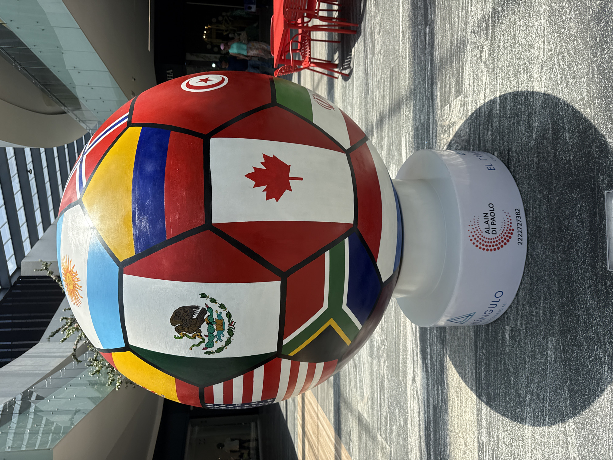 A large soccer ball sculpture is displayed on a white pedestal, likely indoors in a commercial space in Puebla, Mexico. The ball is decorated with national flags. Flags visible include Canada, Mexico, Argentina, Tunisia, and South Africa. The pedestal has text on it, including the name "ALAIN DI PAOLO" and the phone number "2222727382." Some red chairs can be seen in the background. The floor is gray and textured. A shadow cast by the ball and pedestal is visible on the floor.