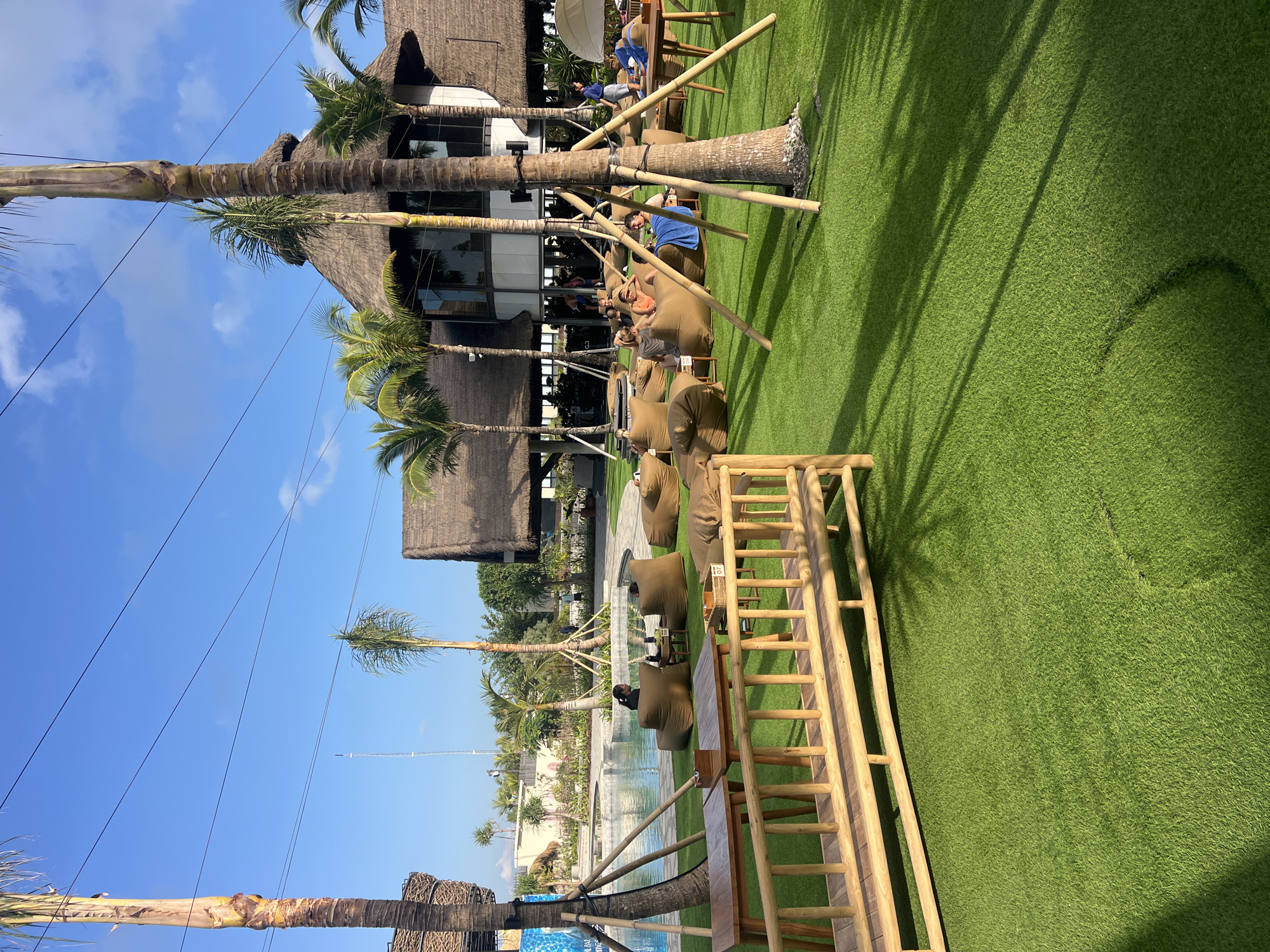 An outdoor scene shows a resort or restaurant with artificial turf lawn in North Kuta, Indonesia. Palm trees with thatched roofs are visible, along with a swimming pool. Several people are seated on beanbag chairs on the lawn, some interacting with each other and others relaxing individually. In the foreground, there are low wooden tables and benches. The sky is clear blue with a few clouds.