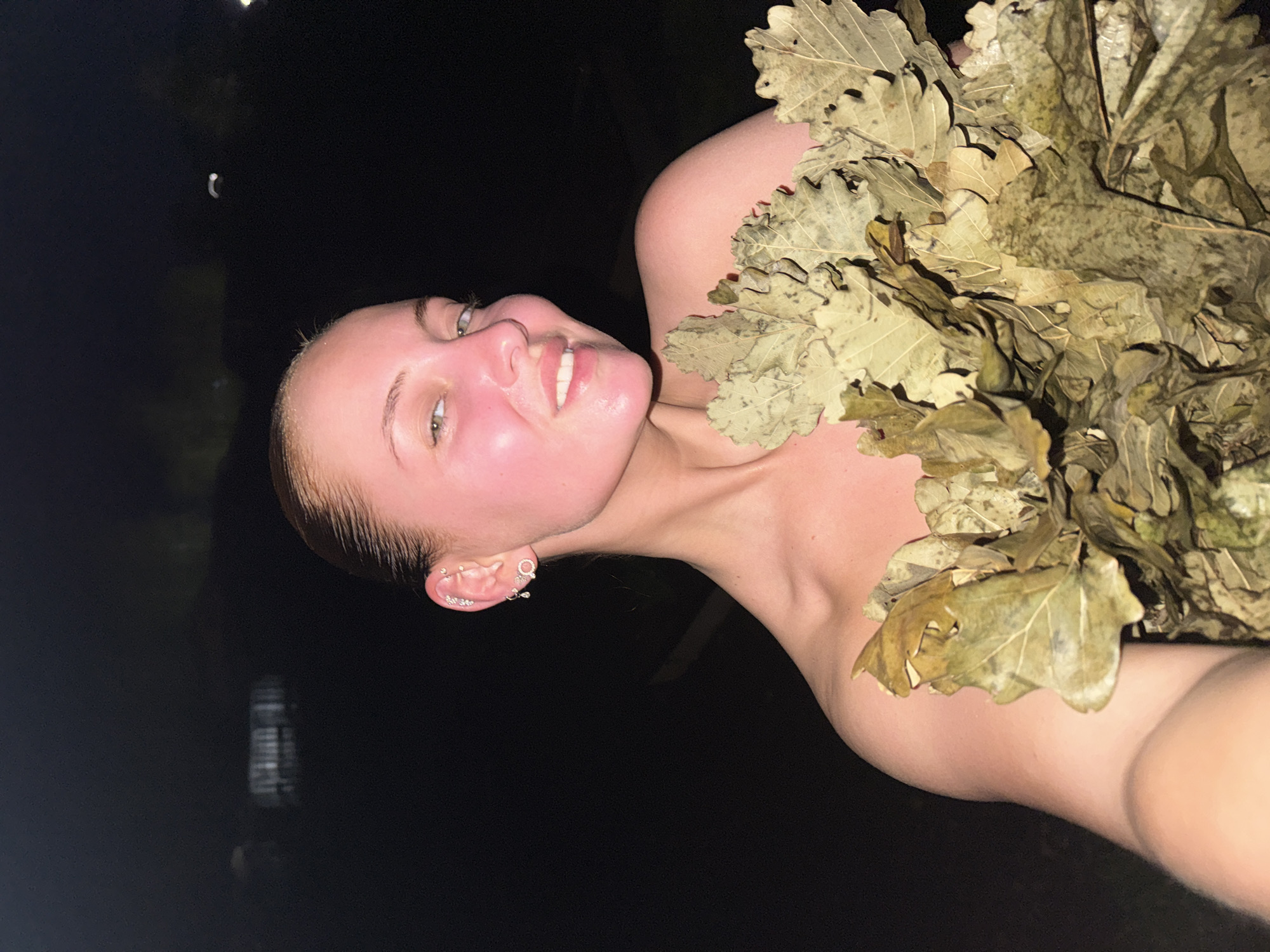 A fair-skinned woman with rosy cheeks is smiling and holding a bundle of dried oak leaves. The background is dark, with what appears to be a body of water visible in the upper left, suggesting a location at night near a lake or river. The woman has multiple earrings in her left ear. It is difficult to identify with certainty the exact location within Chastyye, Russia, based on this image.