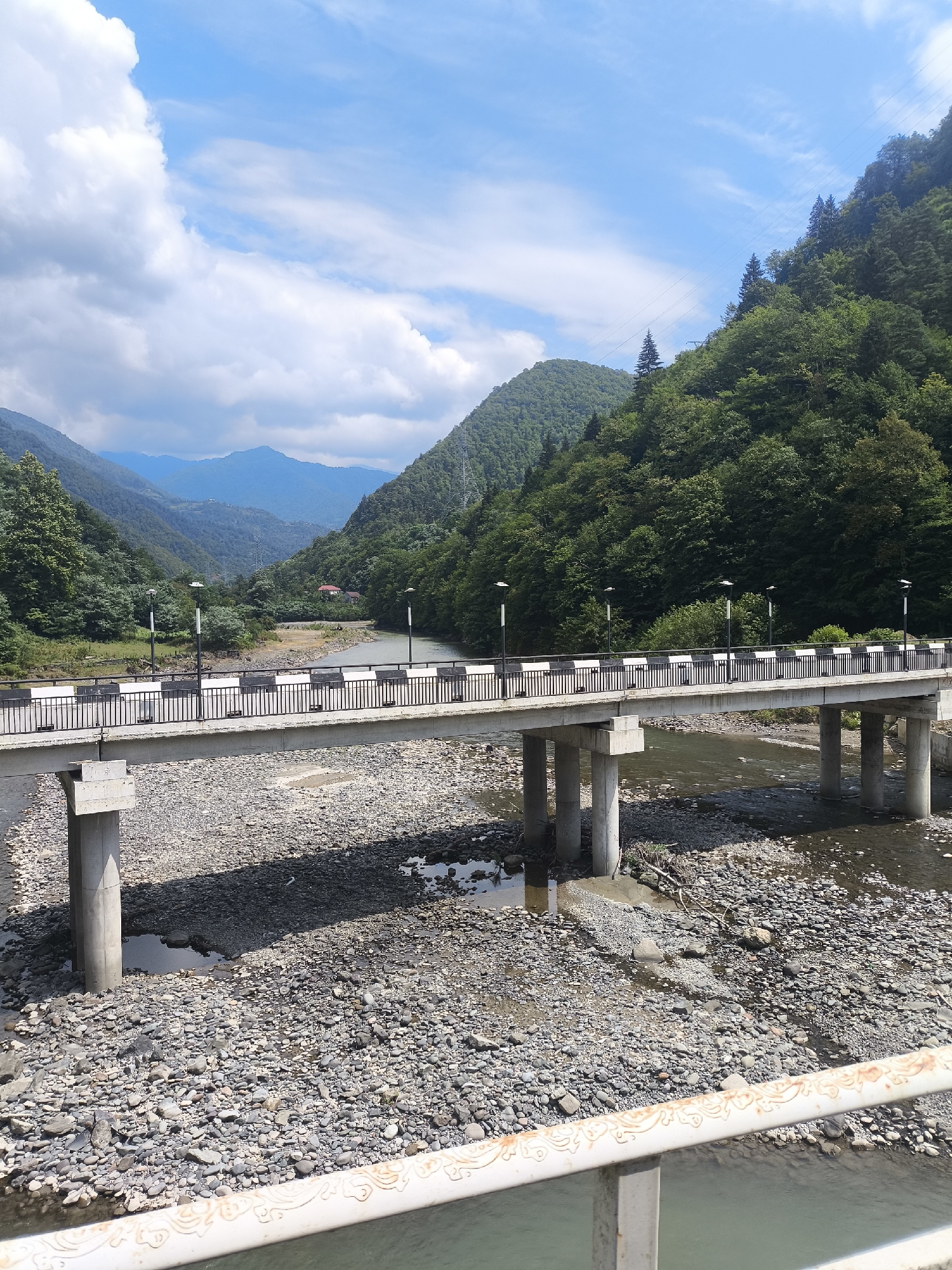 This image shows a bridge crossing a rocky riverbed in the city of Zvare, Georgia. The bridge is made of concrete with a metal railing and lights. The river is shallow and filled with stones. The surrounding landscape is mountainous and covered with lush green trees. The sky is blue with scattered white clouds. There are no people or animals visible in the image.