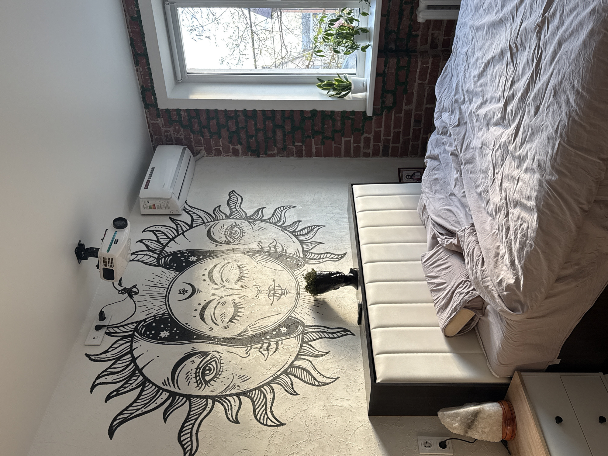 The image captures a distinctive interior room, likely a bedroom or studio apartment, from a high-angle perspective looking downwards. The setting is Rostov-on-Don, Russia. Bright daylight streams through a large window, illuminating the space.
A prominent feature is a large, elaborate mural covering a significant portion of the floor or a large wall area. This mural depicts a celestial theme with three stylized faces resembling suns and moons, each adorned with intricate details like rays, stars, and crescent moons. The artwork is predominantly black on a light, textured surface.
To the right of the mural is a bed with light grey, crumpled bedding, suggesting recent use. Adjacent to the bed is a white, padded bench or headboard. Below the bed, a wooden nightstand holds a glowing Himalayan salt lamp. A wall outlet is visible near the nightstand.
On the opposite side, mounted on a white wall above the mural, is a white projector with its lens and vents visible, connected to a power outlet. Below it, a white portable air conditioner or heater unit, branded "DOMOTEC Previn," rests on the floor.
The wall behind the window and extending towards the bed features a textured surface mimicking red brick, interspersed with green, vine-like artificial foliage. The window itself offers a view of bare tree branches, suggesting a natural outdoor element. Two potted plants, one with green foliage and another with variegated leaves, adorn the white windowsill. A small black pot with a plant is also visible near the padded bench.
No people are present, and no specific activity is taking place. The room appears to be a personal space, comfortably furnished and decorated with a unique, mystical aesthetic. The bright light indicates it is daytime.