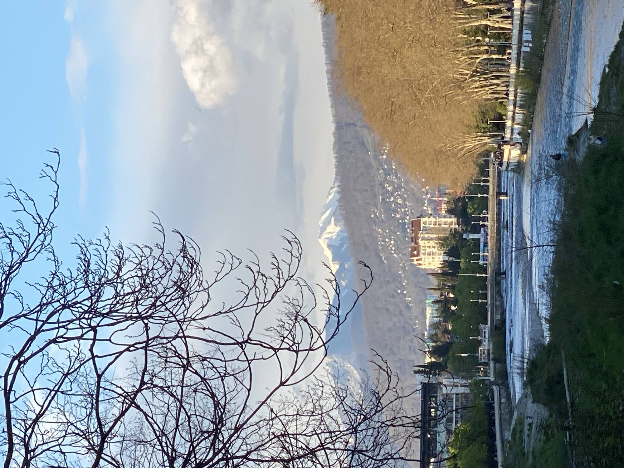 The image depicts a cityscape with a river running through it. On the left, bare tree branches frame the view. In the background, snow-capped mountains rise behind buildings, indicating the city of Sochi, Russia. Along the river, there are lampposts, trees, and buildings. On the river's edge, two people can be seen. The sky is partially cloudy.