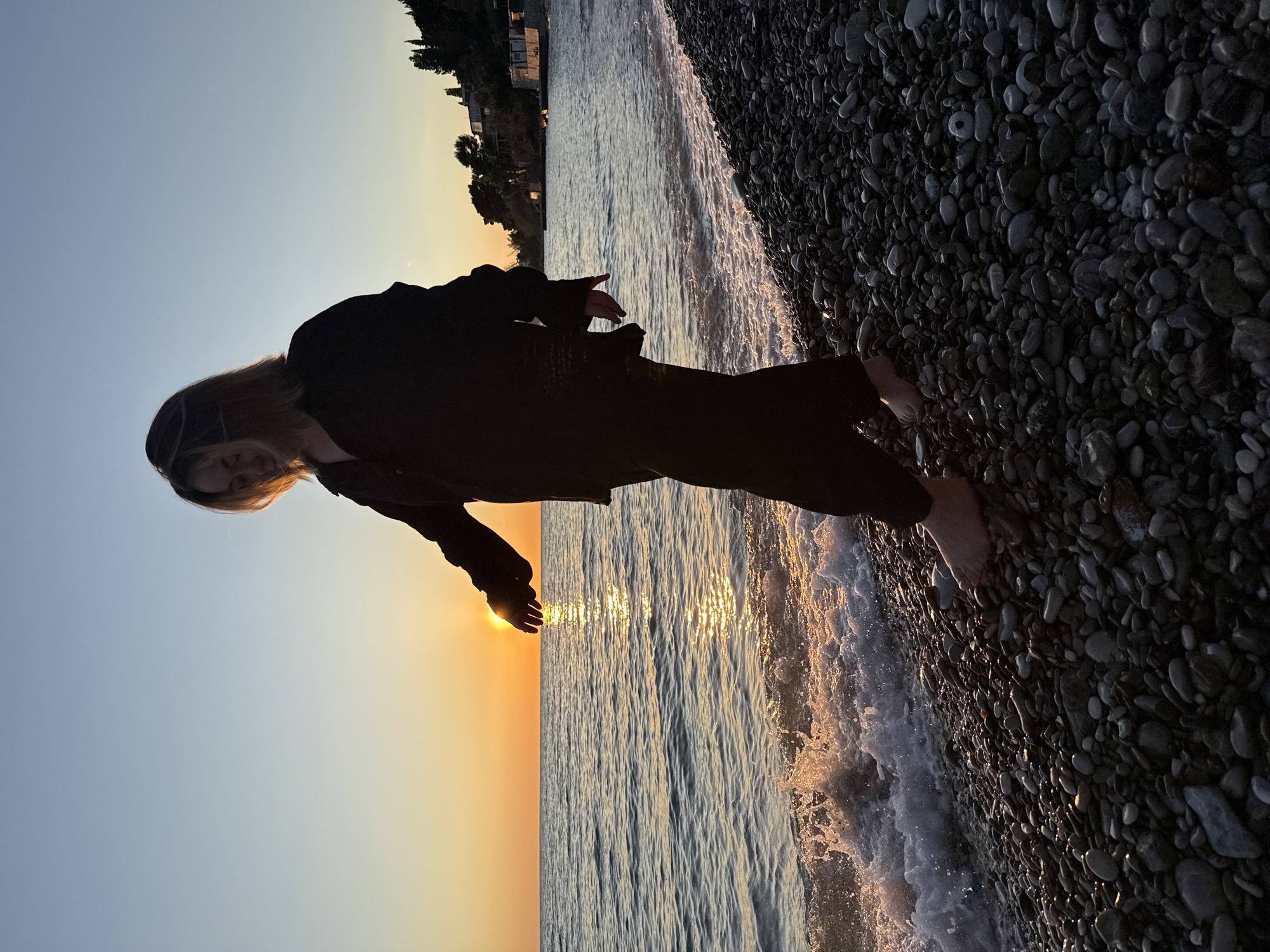 A photograph captures a person standing on a pebble beach in Bar, Montenegro, at sunset. The sky transitions from a clear blue to an orange-tinted horizon. The person is silhouetted, wearing dark clothing and standing barefoot at the edge of the water. Their hand is extended towards the setting sun. The waves are breaking on the pebble beach, and buildings are visible in the distance.
