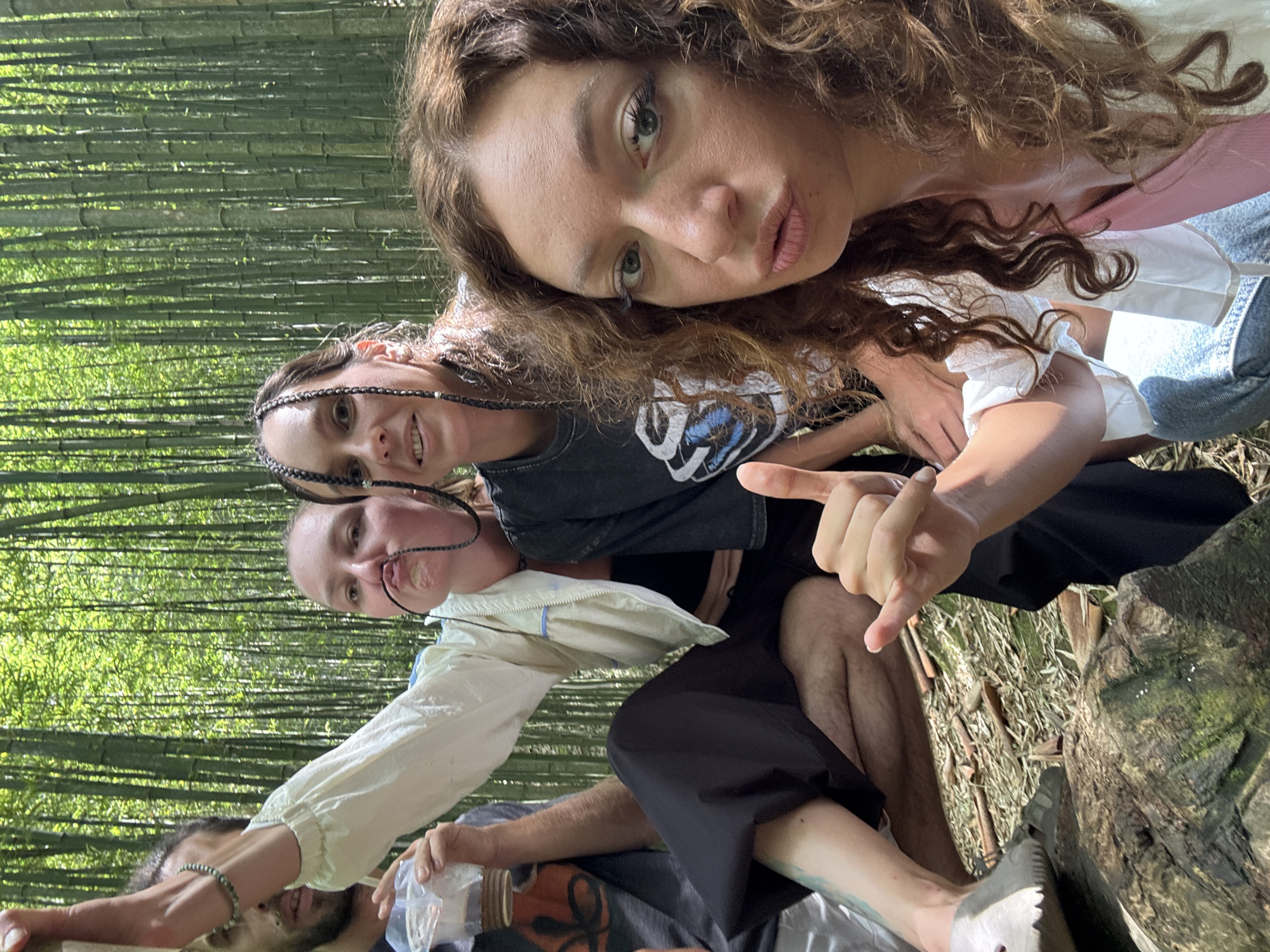 Three young women and a man are photographed at eye level in a bamboo forest, likely located in the region of Púng Luông, Vietnam, given the presence of dense bamboo. The woman at the top of the frame, with curly brown hair, looks into the camera with pursed lips. Below her is a woman with hair braided close to her head, smiling. The third woman, lower down, puckers her lips towards a length of string extending from her mouth. A man with dark hair and a beard is near the bottom of the frame, holding a container of some sort. The environment shows a dense stand of bamboo stalks. The foreground shows dry leaves and debris. The scene suggests a casual gathering or outing.