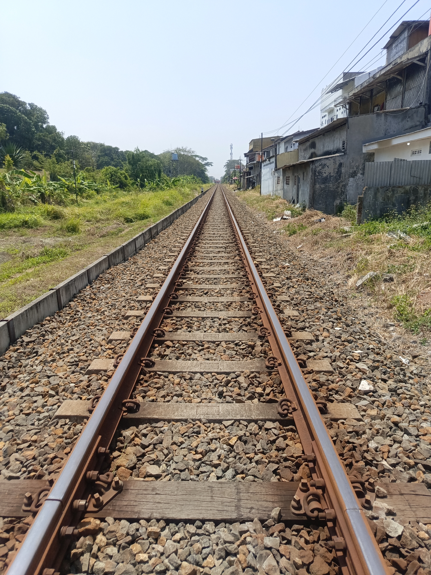 The image depicts a straight railway track receding into the distance. The tracks are made of steel and laid on wooden sleepers, surrounded by gravel. To the left of the tracks, there is a concrete drainage ditch followed by a grassy embankment with trees and vegetation. To the right of the tracks, there are buildings that appear to be residential or commercial, with some signs of wear and tear. Overhead power lines are visible. The sky is clear and blue, suggesting a sunny day. The scene is set in Desa Gunungsimping, Indonesia, as indicated by the context. There are no people visible in the image. The overall impression is of a functional railway line running through a semi-urban or peri-urban area.