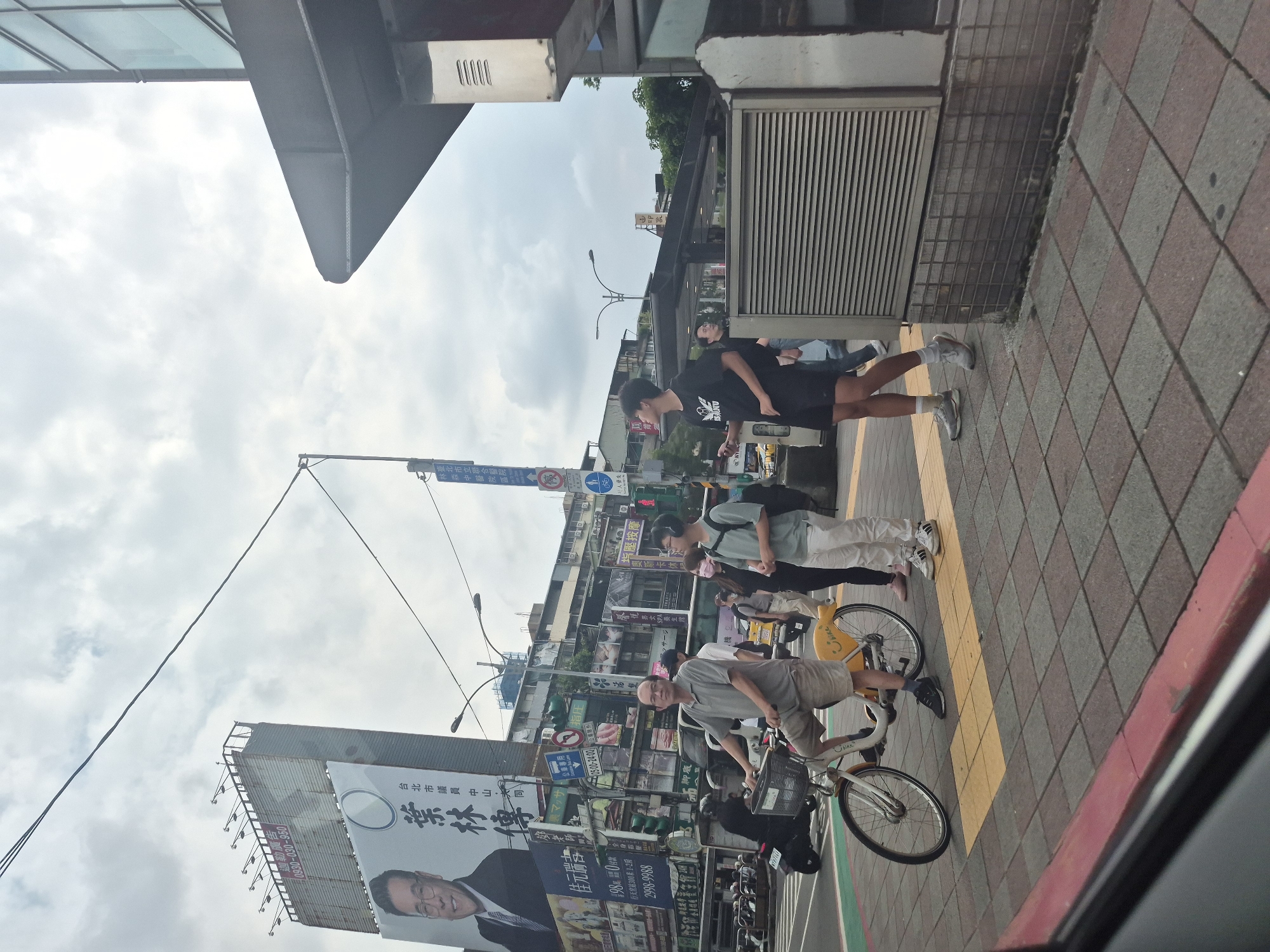 The image depicts a street scene in Taipei, Taiwan, likely an intersection with a pedestrian crossing. In the foreground, a man wearing a grey polo shirt and khaki shorts is seated on a yellow public bicycle. To his right, two young men are standing on the sidewalk. One, in a black t-shirt and shorts, holds a mobile phone. The other, in a light green t-shirt and white pants, wears headphones. A woman in a white t-shirt and black pants, wearing a pink mask, is walking behind the man on the bicycle.
Behind the individuals, the street is busy with traffic, including motorcycles and cars. Billboards are prominently displayed on buildings along the street. One large billboard features a portrait of a man in a suit and text in Chinese characters. Traffic lights and street signs are visible. The environment suggests an urban setting with a cloudy sky. The sidewalk is paved with checkered tiles, and a red curb is visible at the edge of the road.