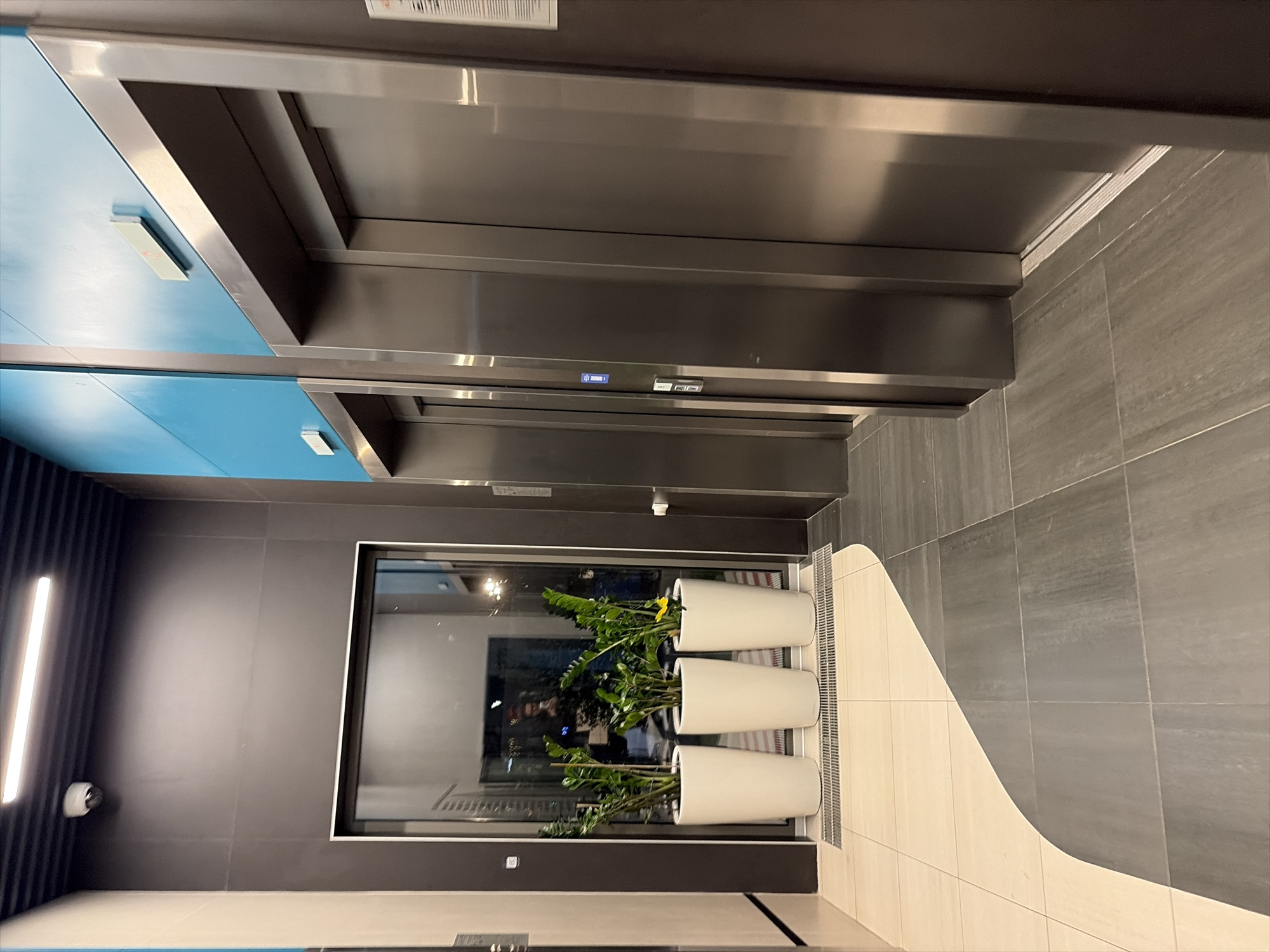 A modern, indoor elevator lobby in Moscow, Russia, features two polished stainless steel elevator doors on the right. Above the doors, a teal blue wall is punctuated by a light-colored rectangular indicator. To the left of the elevators, a large glass window reveals a dimly lit interior with what appear to be stair railings, suggesting the presence of a staircase. In front of this window, three tall, white cylindrical planters hold lush green plants, with one plant displaying a single yellow flower. The floor is a mosaic of light beige and dark gray square tiles, arranged in a flowing, abstract pattern. A security camera is mounted on the black ceiling with linear lighting. The scene is devoid of any visible people or ongoing activity.