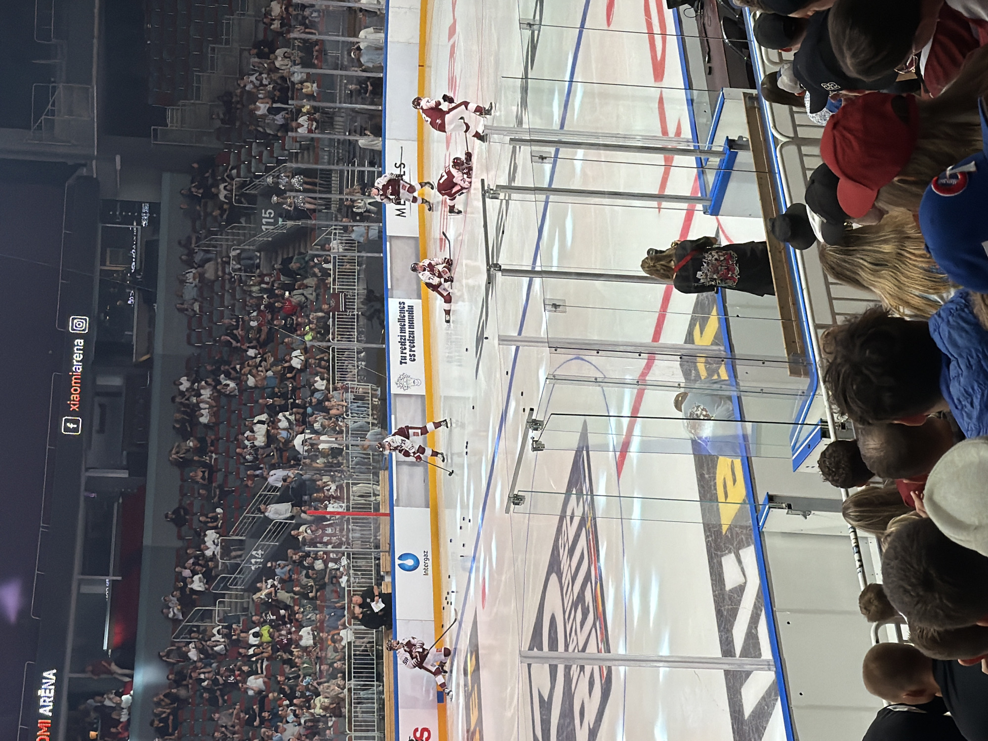 The image depicts an ice hockey game taking place inside the Arena Riga, located in Riga, Latvia. The arena is filled with spectators seated in tiered stands, some of whom are visible in the foreground and background. On the ice, several hockey players in red and white uniforms are engaged in play, with pucks scattered across the surface. The arena features advertising boards along the perimeter of the ice, with logos such as "Intergaz" and "Xiaomi Arena" prominently displayed. The lighting suggests it is an evening event. A photographer, identifiable by her camera and black attire, is positioned behind a glass barrier, capturing the action on the ice.