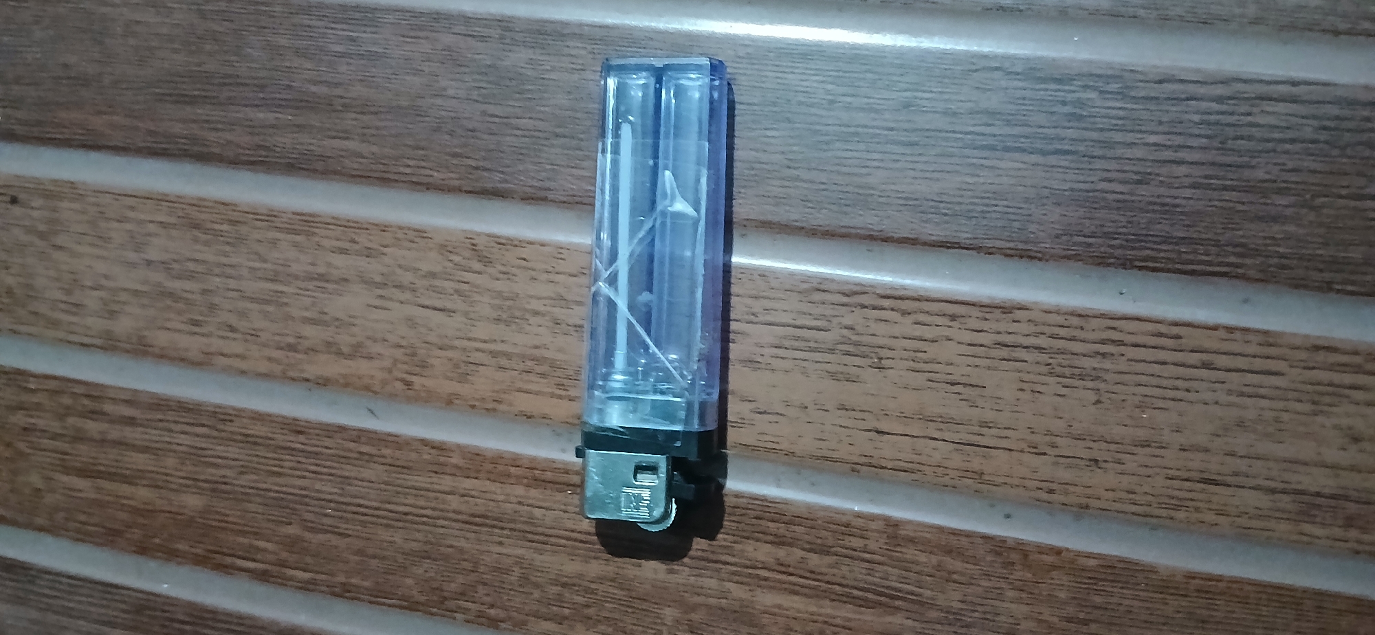 A clear plastic lighter, lying horizontally, is positioned on a brown wooden surface with distinct vertical grooves. The lighter has a metallic striking mechanism on the left and a transparent body containing a purple gas chamber and internal metal components. The background surface exhibits a wood grain pattern with light-colored lines running vertically, suggesting a table or a similar furniture item. There are no people or other objects visible in the scene. The image appears to be taken in Sekarputih, Indonesia, though no specific landmarks or contextual clues within the image itself confirm this location.