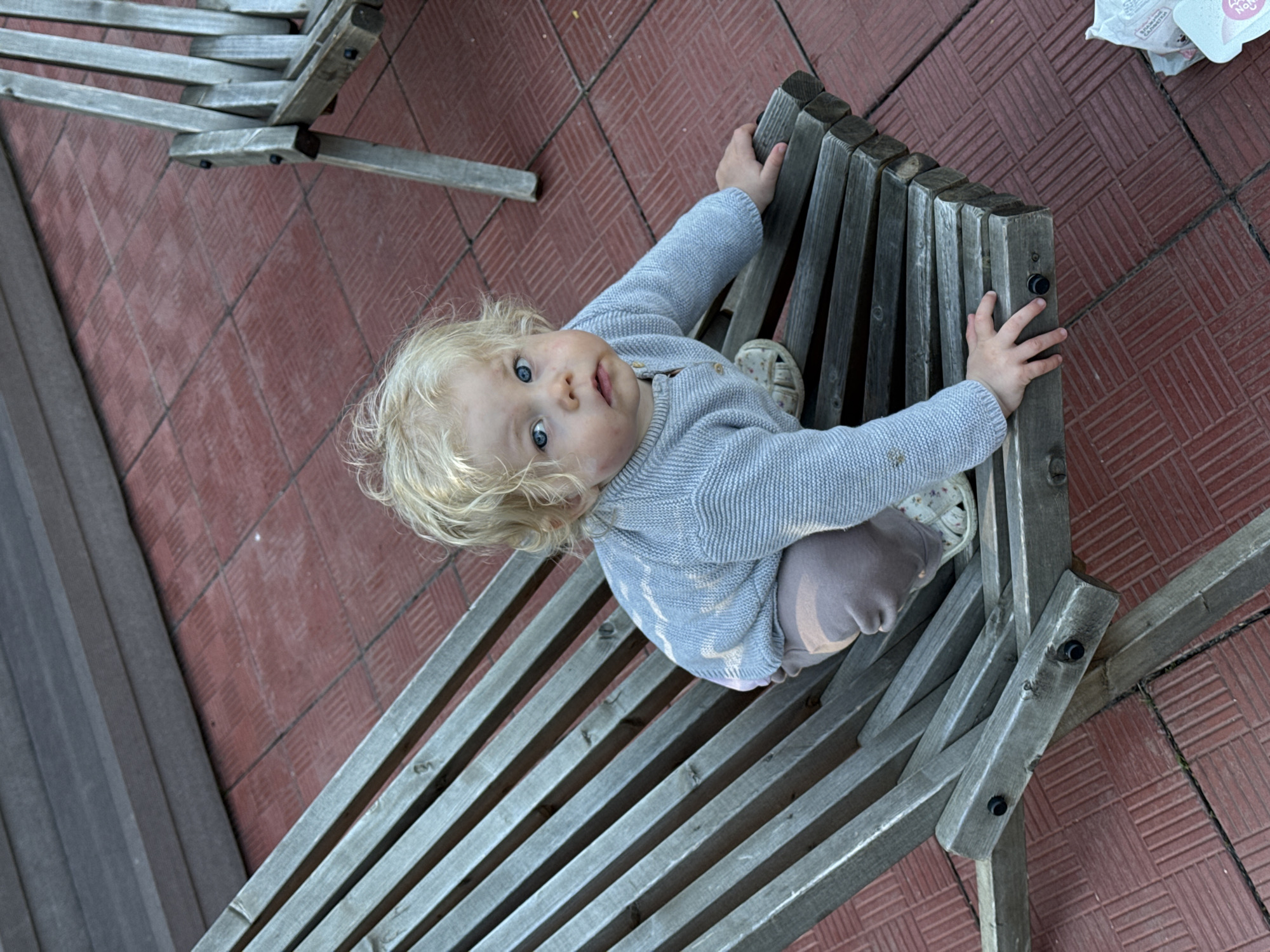 A blonde child with blue eyes, approximately one to two years old, wearing a light grey knitted sweater, light brown pants, and white sandals, is positioned on a weathered, grey wooden lounge chair. The child's hands are gripping the wooden slats of the chair, and the child's gaze is directed upwards towards the camera. Minor smudges are visible on the child's face and sweater. The lounge chair is composed of parallel wooden slats connected by dark fasteners. The ground beneath is covered with reddish-brown square tiles arranged in a patterned layout. In the upper left background, a blurry wooden structure, possibly a wall or elevated platform, is visible. To the right, a portion of another identical weathered wooden lounge chair is present. In the lower right corner, a white plastic bag and a light blue item with non-English text are visible on the tiled surface. The scene is outdoors, illuminated by natural light in Копнино, Russia.