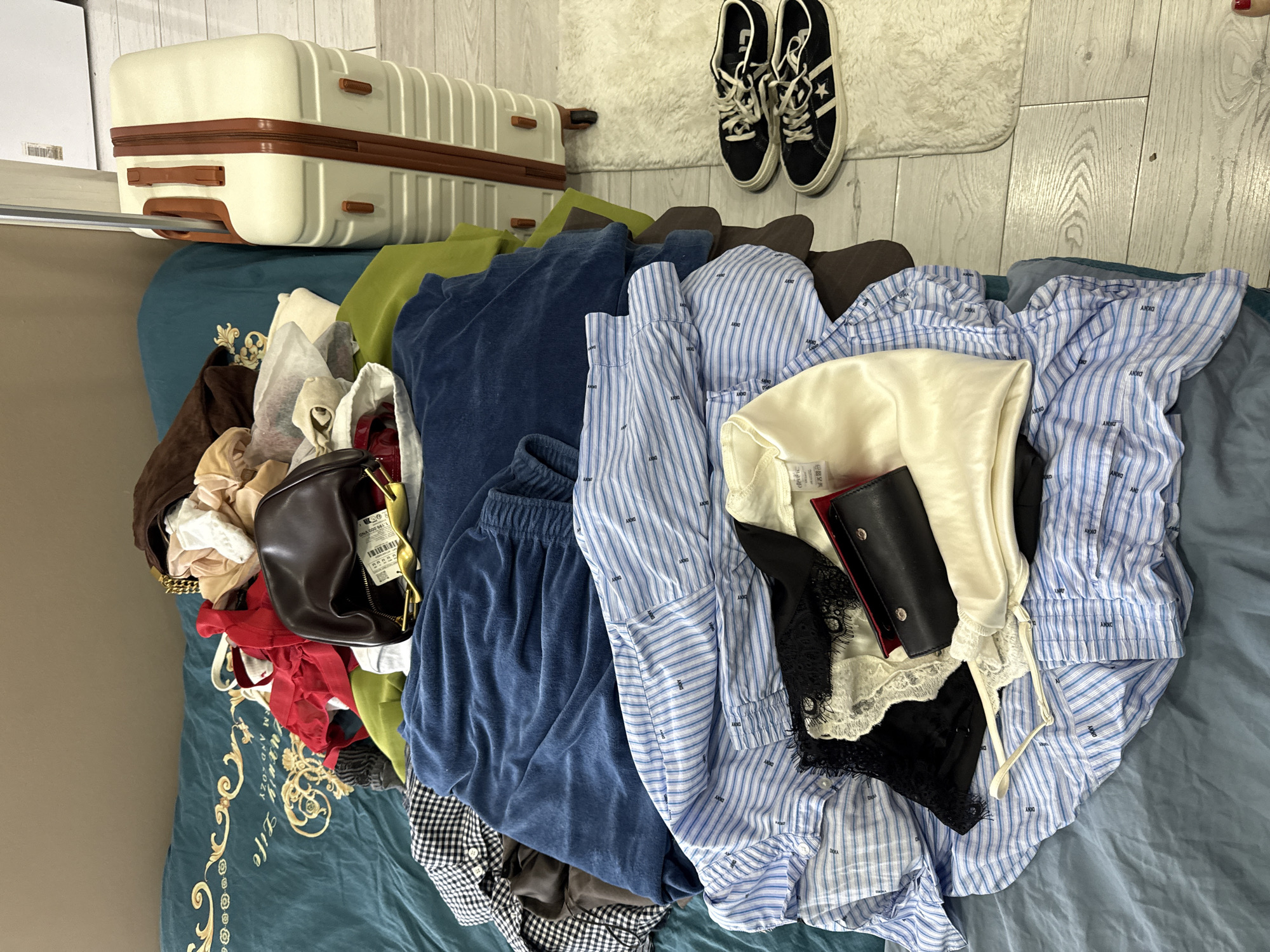 The scene is a cluttered indoor space, likely a bedroom, with a bed covered in a teal-patterned comforter. A light-colored suitcase with brown accents sits on the bed. Adjacent to the suitcase, on a light-colored wooden floor with a white rug, are a pair of black sneakers. The bed is piled with a variety of clothing items in various colors and textures, including a blue velvet tracksuit, striped shirts, a cream-colored satin top, and black lace lingerie. A brown leather handbag and a black wallet are also among the items. There are no people visible in the image. No indications of Sochi, Russia, are present in the image.