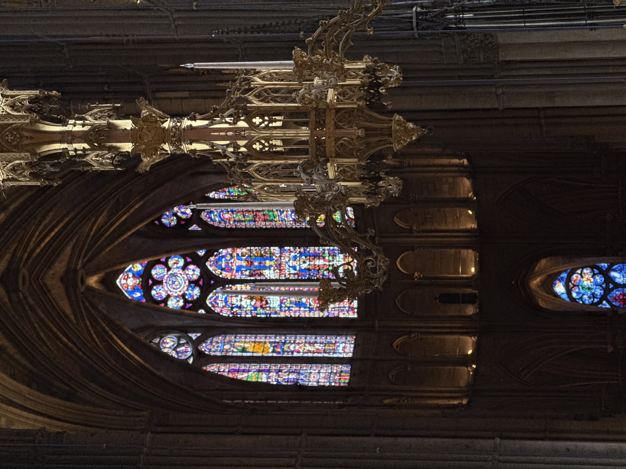 The image captures a detailed view inside a grand Gothic cathedral, likely Reims Cathedral given the provided context. The perspective is an upward, interior shot, dominated by an exceptionally ornate, gilded metal chandelier or light fixture that hangs prominently in the upper center of the frame. This elaborate piece features intricate gothic tracery, spires, and delicate scrollwork, characteristic of medieval ecclesiastical design. Its metallic surface reflects some light, highlighting its sculptural details.
Below and to the left of the chandelier, a large, multi-part stained glass window illuminates the interior with vibrant blues, reds, greens, and yellows, depicting religious scenes or patterns. This window is set within a deep, arched stone opening, revealing the massive scale of the architecture. Further to the right, another, smaller circular stained glass window with a distinct rosette pattern is visible, also glowing with color. The surrounding architectural elements are composed of dark stone, showcasing vaulted structures, arches, and possibly triforium or clerestory levels in the background, subtly lit by unseen sources or ambient light.
The scene conveys a sense of awe and historical grandeur, typical of European cathedrals. The light filtering through the stained glass suggests it is daytime, although the overall interior is dimly lit, enhancing the dramatic effect. There are no people visible, and no specific activity or event is taking place beyond the static presence of the architecture and its decorative elements. No visible text is present in the image.