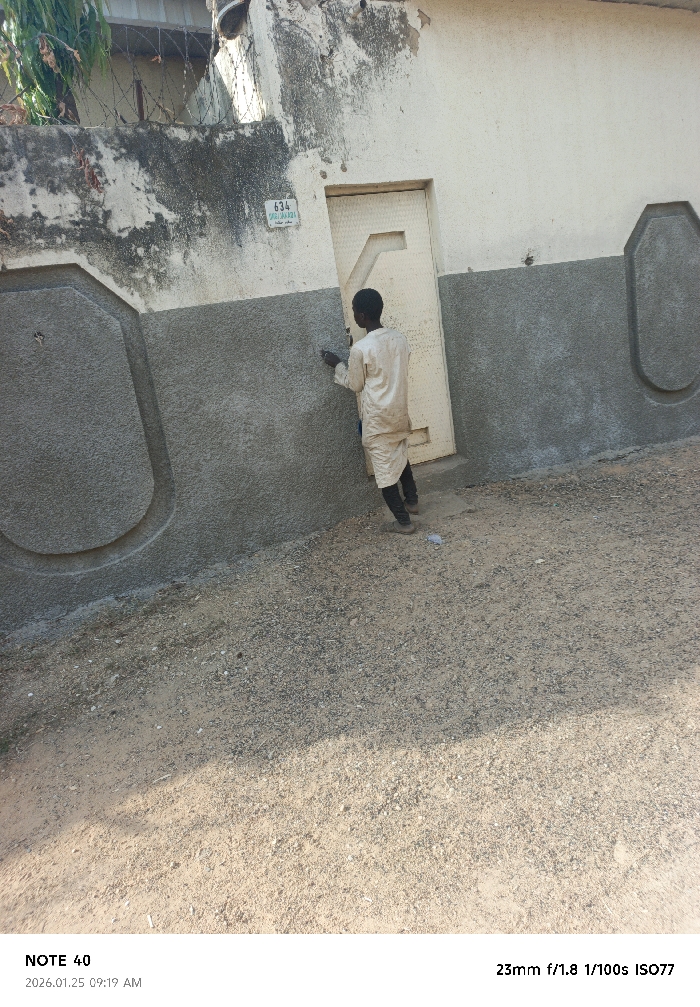 In Panshekara, Nigeria, an outdoor daytime scene captures a single individual, appearing male, with dark skin, positioned with his back mostly to the camera. He wears a light-colored, long-sleeved tunic over dark trousers and shoes. His right hand holds a small, dark object against the lower portion of a building wall. The wall is two-toned: an upper, weathered off-white section, and a lower, textured dark grey section. A light green rectangular sign marked "634 GABA MALAGA" is affixed to the upper wall, positioned above a slightly ajar cream-colored door featuring a geometric pattern. The lower dark grey wall section includes two prominent, light grey, oval-shaped decorative panels. Above the wall to the left, a wire fence topped with barbed wire is visible, along with some green foliage. The ground consists of light brown dirt and dark gravel, with a shadow partially obscuring the lower right foreground.