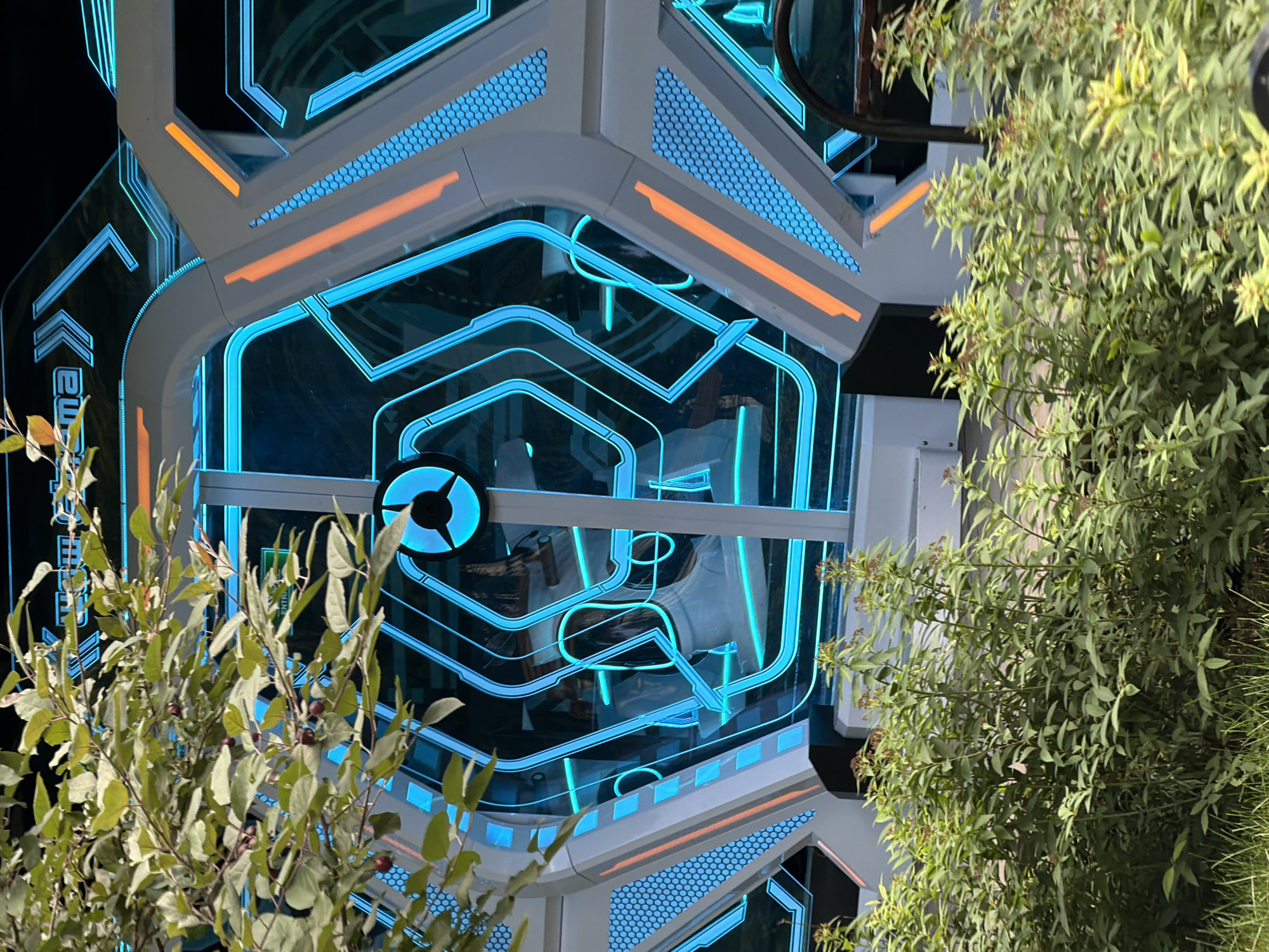 This is a medium shot, eye-level photograph of the entrance to an amusement park ride or attraction, likely in Perm, Russia. The structure features a futuristic, hexagonal design with bright blue neon-like lighting outlining geometric shapes. Orange accent stripes are visible on the white, angular exterior. Through the transparent panels of the entrance, interior seating or capsule-like structures can be seen, illuminated by the blue light. In the foreground, out-of-focus green foliage and grass obscure the lower portion of the scene. The overall impression is one of a modern, technologically advanced attraction.
