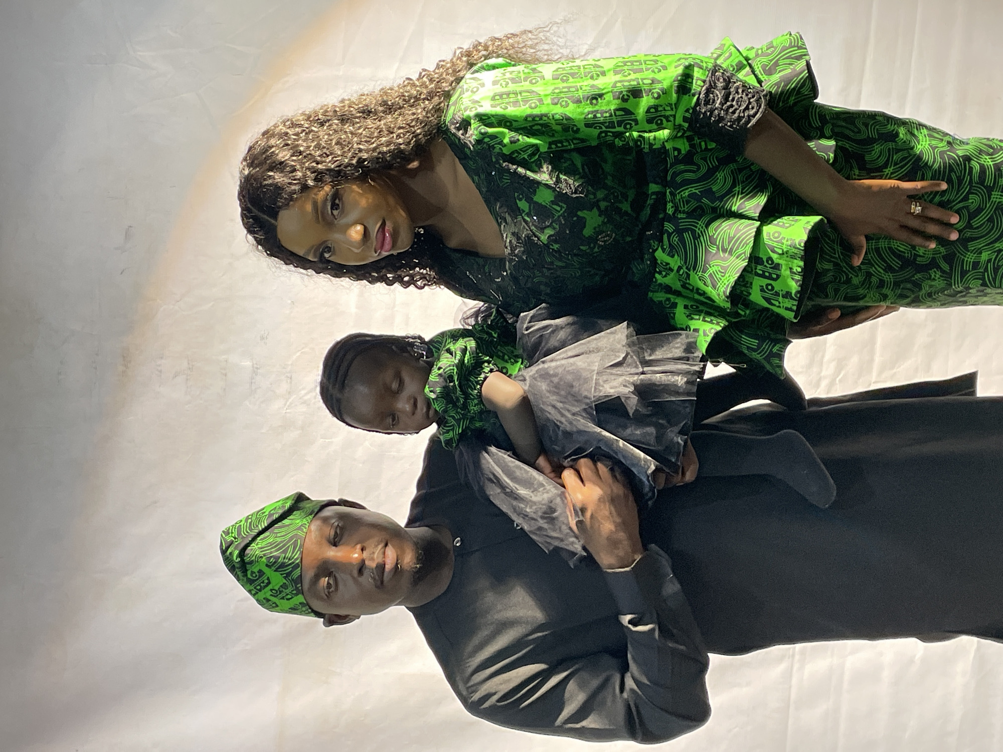 A family of three is posing for a photo. The father, wearing a black shirt and green patterned hat, is holding a child wearing a green and black patterned dress with a black tulle skirt. The mother, standing beside them, is wearing a green and black patterned dress with black lace details. The mother has long curly brown hair. The background appears to be a white or light-colored fabric backdrop. The location cannot be determined to be Jos, Nigeria, from the image.