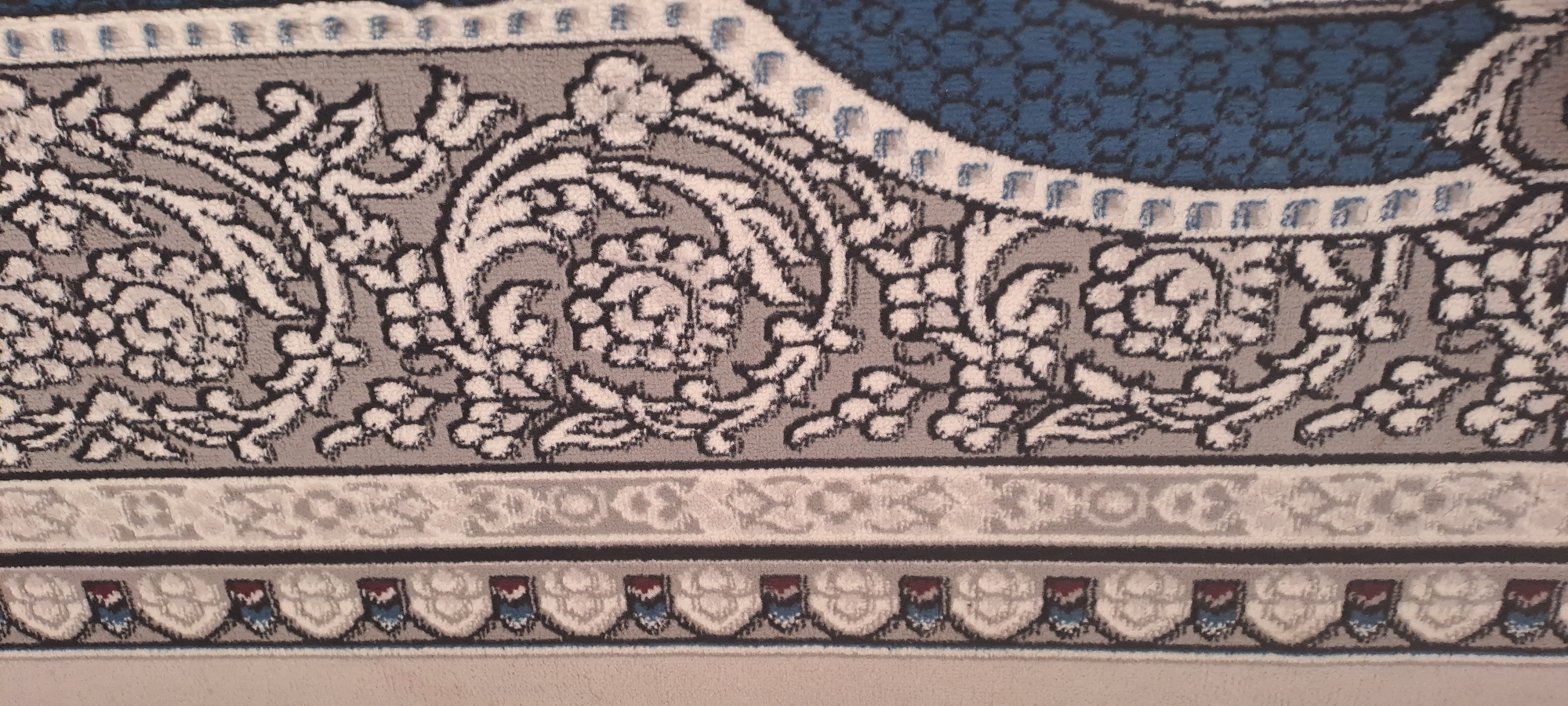 The image displays a close-up view of a rug with an intricate pattern. The rug features borders with repeating motifs in shades of white, gray, blue, and red. The main body of the rug exhibits a large floral or paisley design in black and white against a gray background. A portion of a dark blue textured area is visible on the right side of the image, suggesting a larger patterned design. The texture of the rug indicates it is likely made of soft fibers. The lighting is neutral, and there are no people or any contextual elements to suggest a specific location within Andijan City, Uzbekistan.