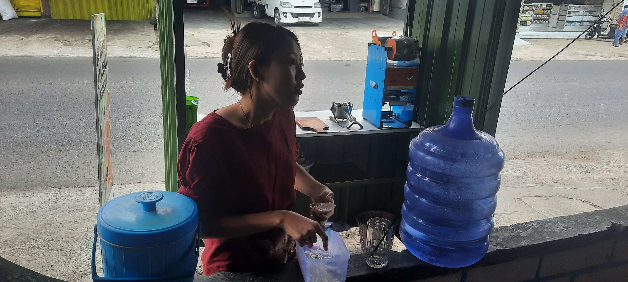 The image shows a street-side vendor in Selopuro, Indonesia. A woman wearing a dark red shirt is preparing drinks. She has her hair pulled back with a clip and is looking to her right. In front of her is a blue plastic cooler and a clear plastic container with shaved ice and a scoop. To her right, there is a glass, a trash can, and a large blue water jug. Behind her, there is a blue drink sealing machine and a wooden board. The background shows a street with a white truck and a building with shelves visible. The scene is set on a paved surface with a grey road in the background.