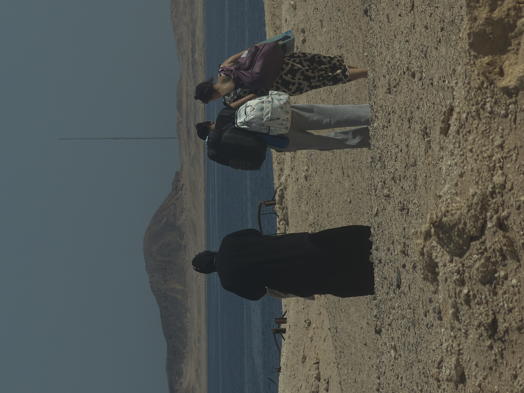 The image depicts a group of three individuals standing on a dry, sandy terrain near a body of water, possibly the Red Sea in Sharm El Sheikh, Egypt. A person wearing a long, dark robe, possibly religious attire, stands facing away from the camera towards the water and a distant, arid landscape with a mountain. To their right, two other individuals, appearing to be women, are also facing away from the camera. One is wearing a backpack and the other carries a large purple bag over her shoulder. The environment is characterized by sparse vegetation and a clear, hazy sky. In the background, a tall, thin structure, resembling a radio tower or lighthouse, is visible against the horizon. The sandy ground is littered with small rocks and debris.