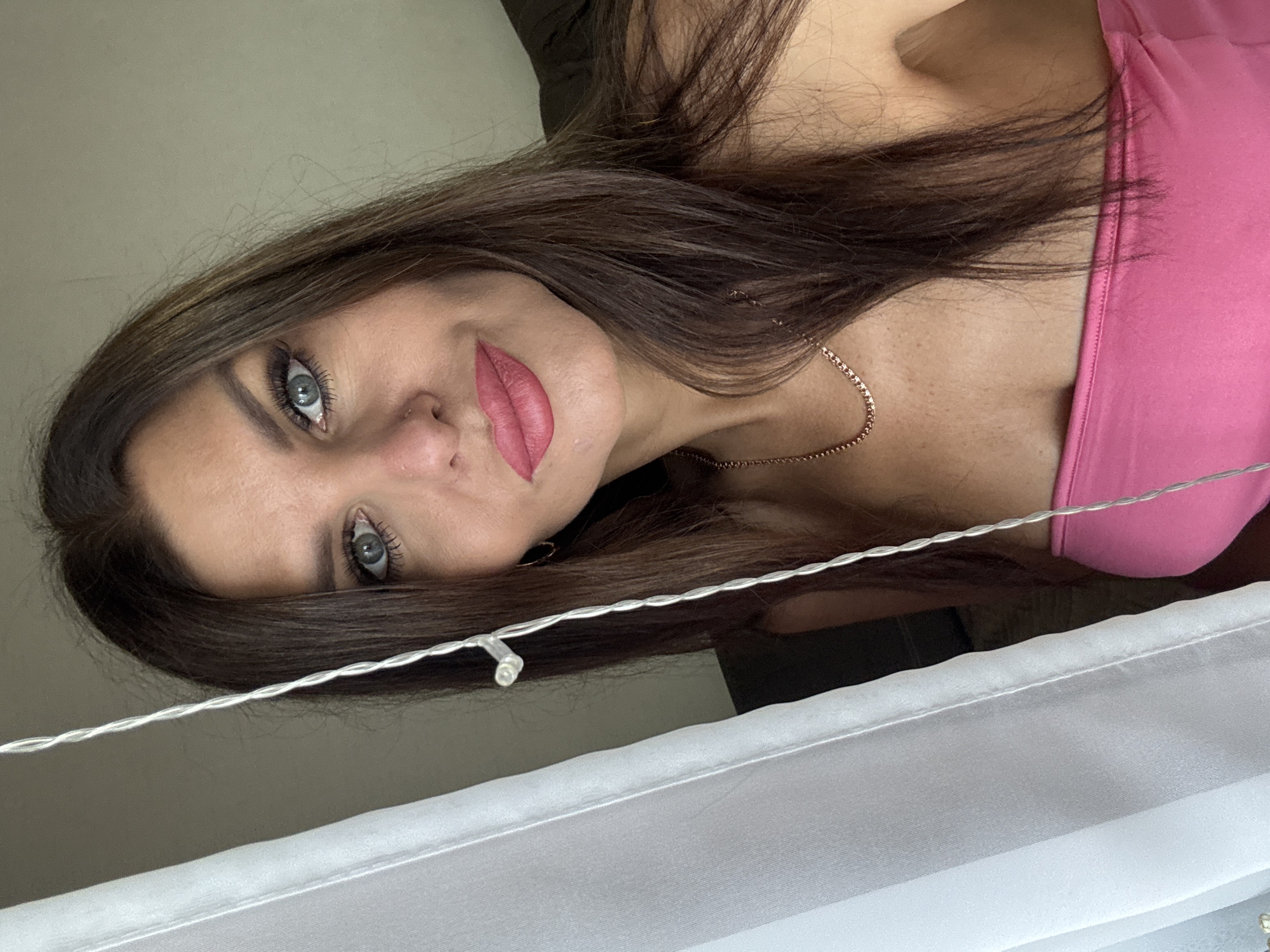 The image shows a woman with long, dark brown hair and blue eyes. She is wearing a pink strapless top and a gold necklace. She has makeup on, including pink lipstick and winged eyeliner. She is looking at the camera and smiling slightly. The background is blurred, but appears to be an indoor setting with a white curtain to the left and a dark sofa to the right. There are also some fairy lights hanging in the foreground. The image is a selfie taken from a medium close-up perspective. No specific location in Chaikovsky, Russia, can be identified.