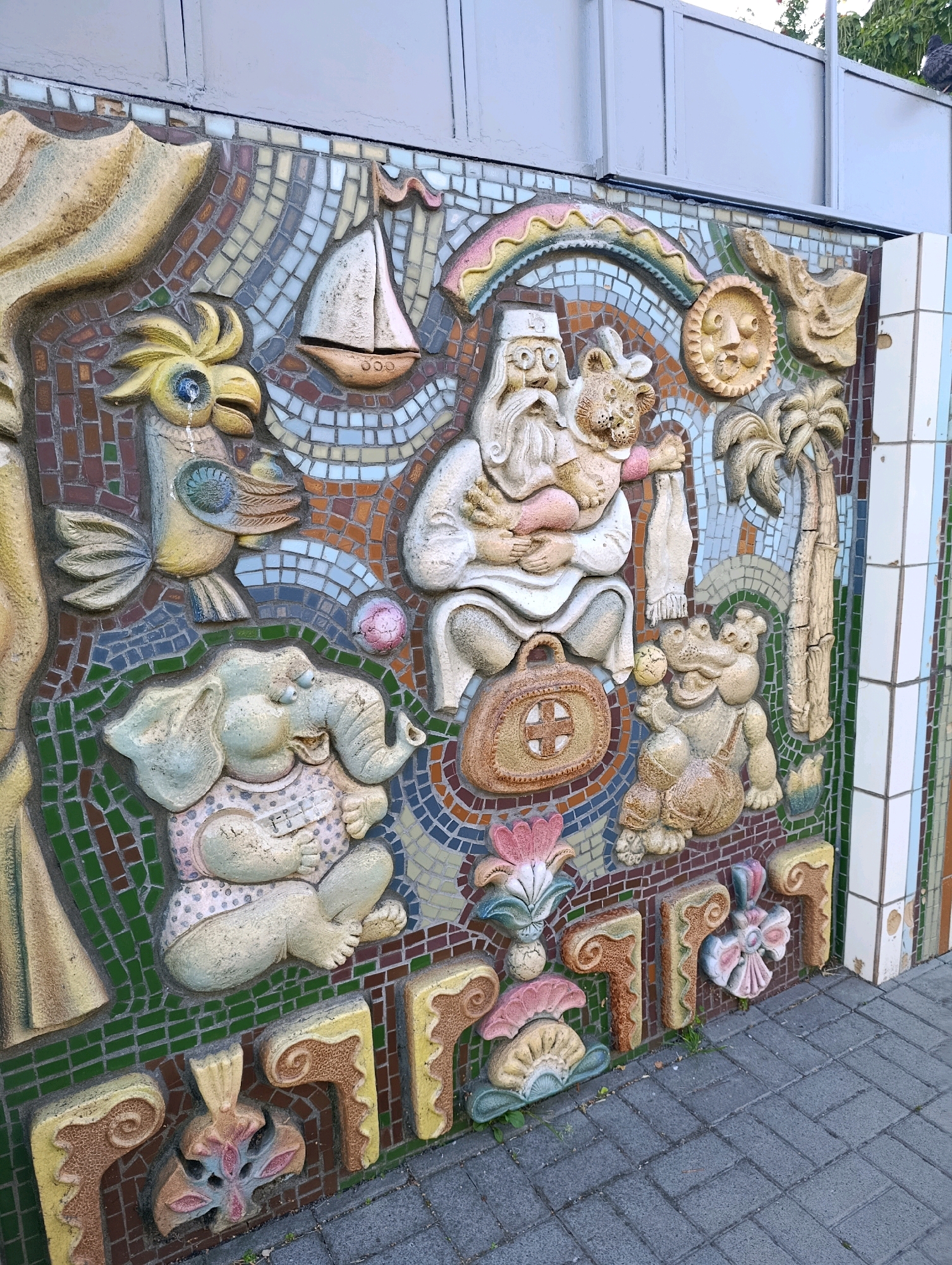 The image depicts a colorful mosaic mural on an exterior wall, likely in Cherkasy, Ukraine, given the context. The mural is highly detailed and features numerous sculpted figures embedded within a mosaic of small ceramic tiles. Prominently visible are an elephant wearing a polka-dot shirt, a bird resembling a parrot with a blue eye, an elderly man with a long white beard wearing a medical-style hat and holding a cat-like creature, and a crocodile-like figure. Other elements include a sailboat, a sun with a face, a palm tree, and various decorative floral and abstract shapes. The mural is affixed to a wall that is partially obscured by a metal fence or partition at the top. The lower portion of the mural transitions to decorative ceramic tiles and is situated above a paved sidewalk made of gray bricks. The overall scene suggests an outdoor public art installation.