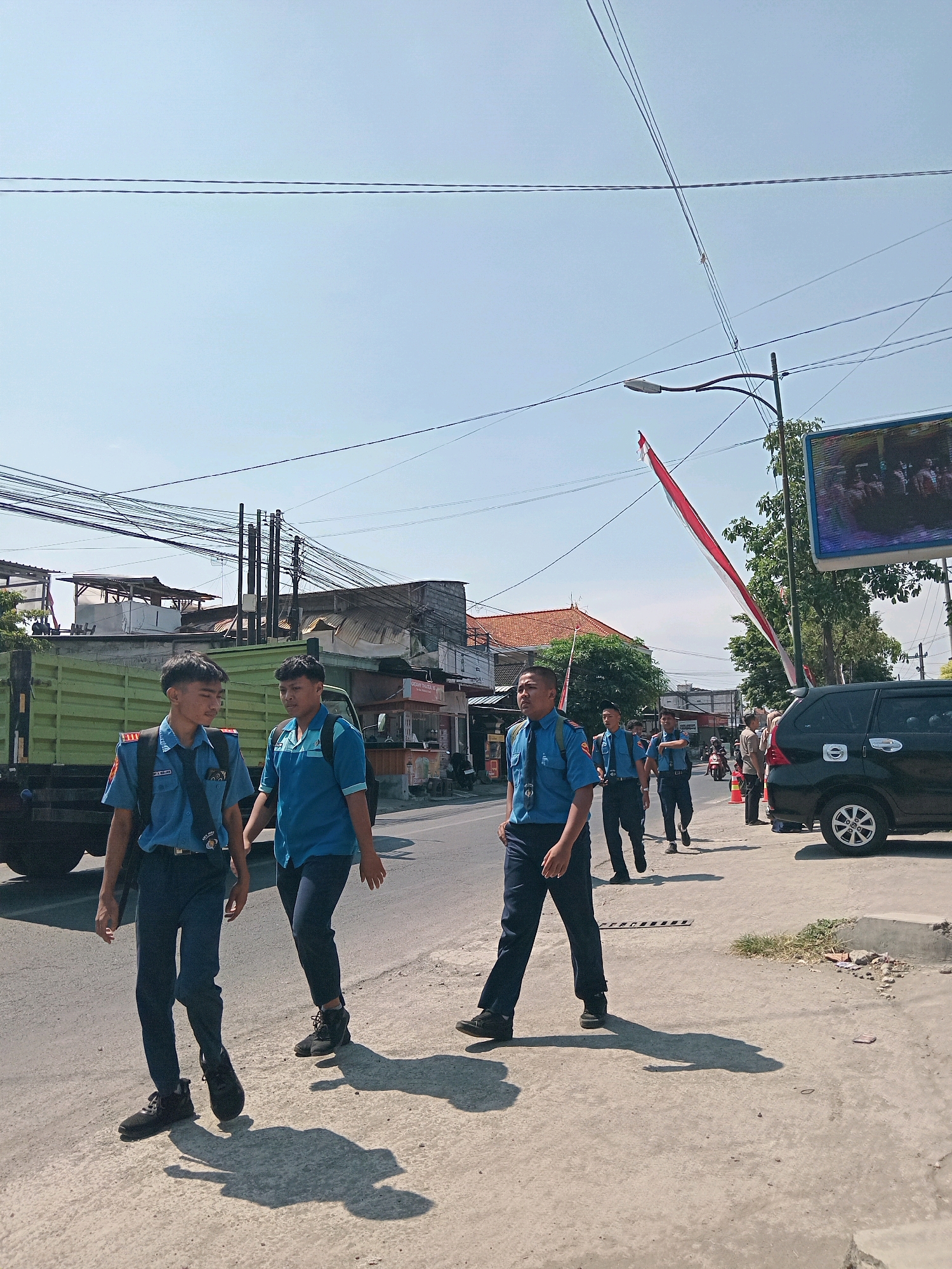 A group of young men in matching blue uniforms walk along a street in Jepun, Indonesia. The men appear to be students or part of a youth organization. They are wearing blue shirts with badges and dark pants. Some have backpacks. They are walking on a paved sidewalk next to a road. In the background, there are buildings, a green truck, and a black SUV. A large Indonesian flag, red and white, is visible hanging from a pole. The sky is clear and blue, indicating a sunny day. The scene appears to be a street in a moderately developed urban or suburban area.