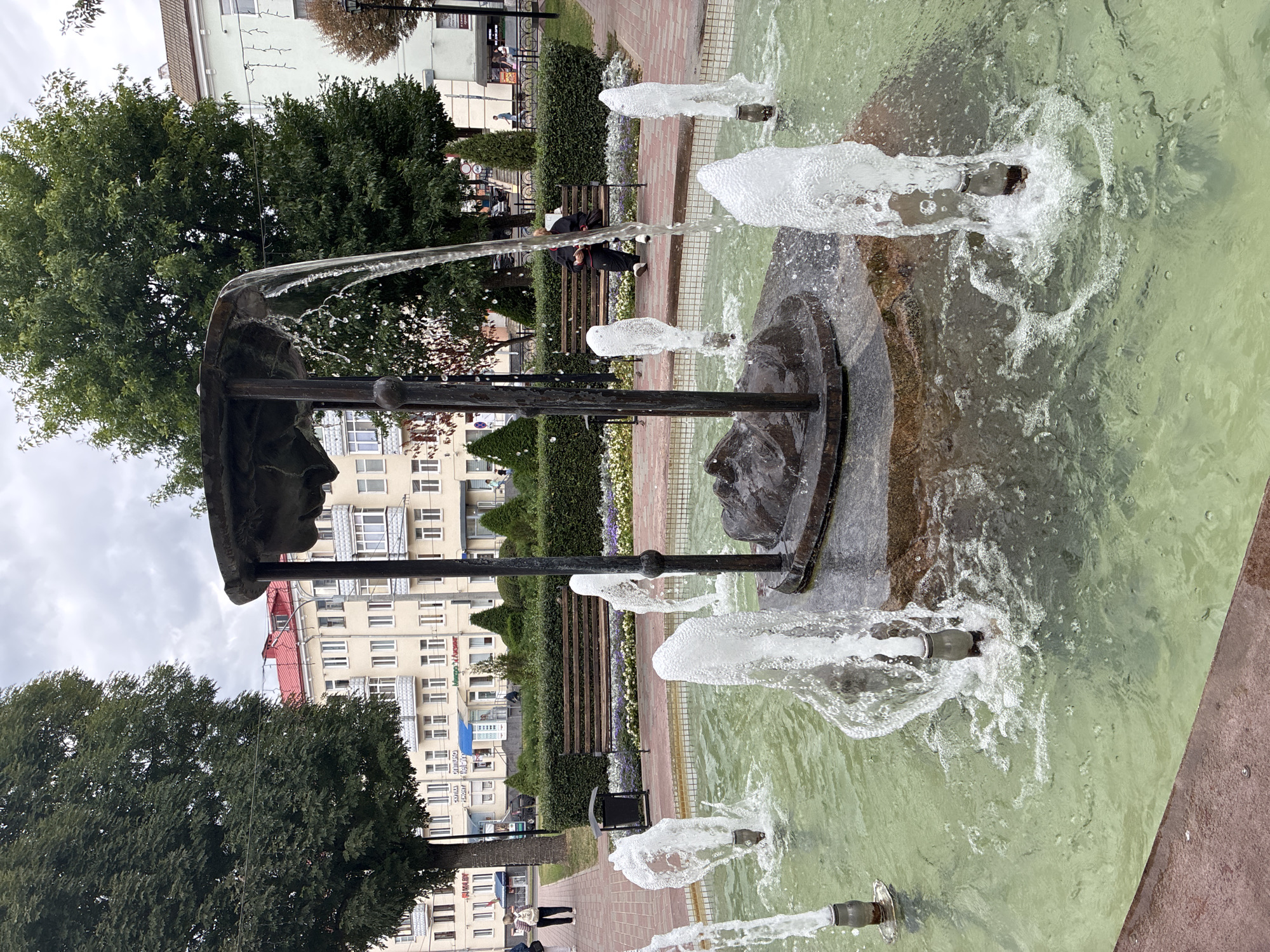 This is a medium-shot, eye-level, outdoor photograph of a fountain in a city park, possibly in Mahilyow, Belarus. The fountain has a central bronze sculpture of what appears to be a sleeping or resting figure on a stone base, surrounded by several spouting jets of water. The water in the fountain basin is a murky green color. In the background, there are trees, a paved walkway, park benches, and several multi-story buildings with red roofs. The sky is overcast. One person is visible sitting on a park bench reading a book. Another person is walking away from the camera on the walkway. The overall impression is of a quiet urban park on a cloudy day.