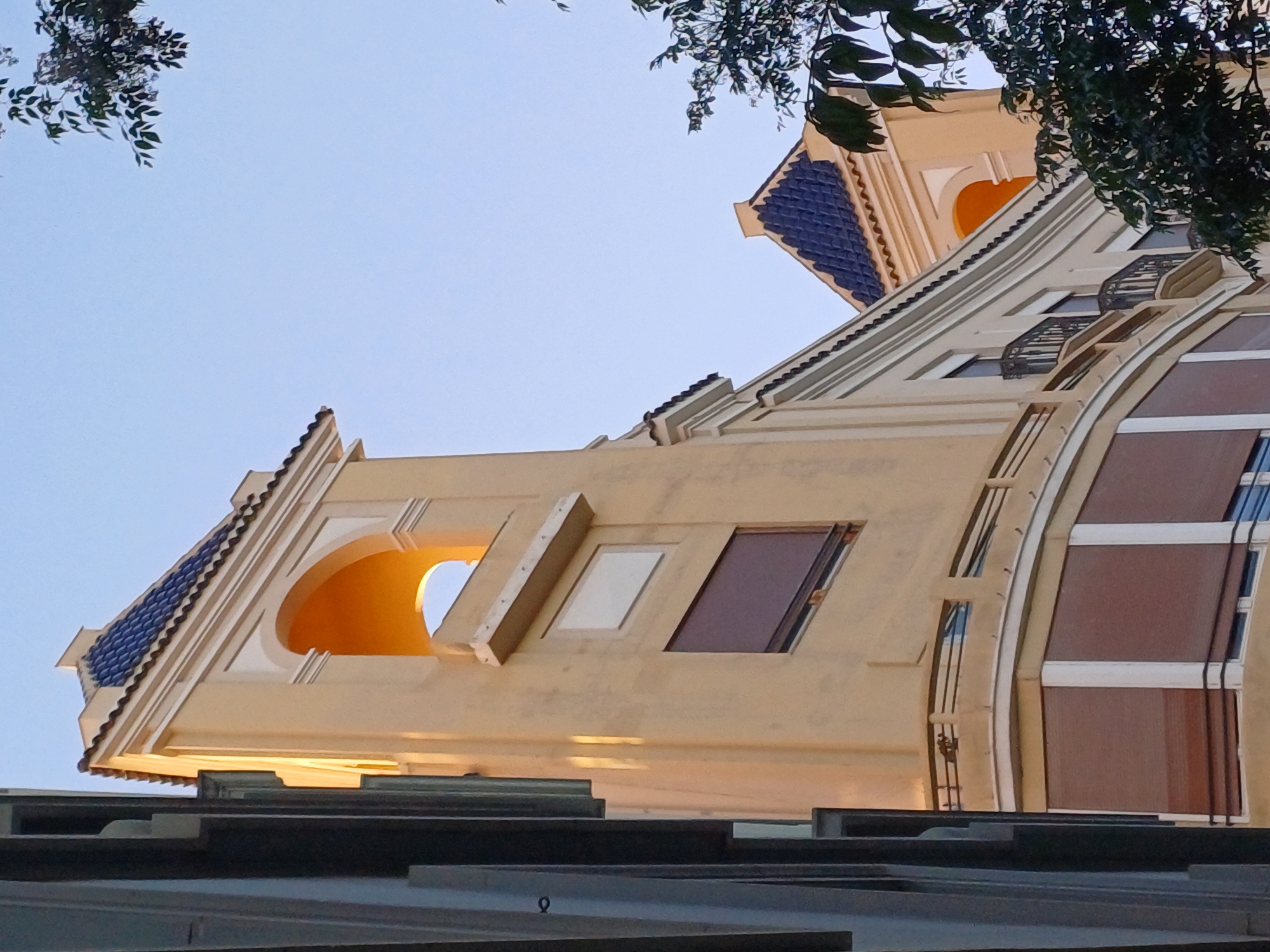 A low-angle, slightly tilted view presents the facade of a building in Valencia, Spain. The architectural design features a beige color palette, contrasted with a blue-tiled roof. There are several window designs, including an arched opening that reveals an orange interior, a rectangular opening with a light-colored surface visible inside, a narrow rectangular window with a brown or dark interior, and an arched window structure with clear panes. The architectural elements are framed by tree branches at the top of the image, set against a light blue sky.