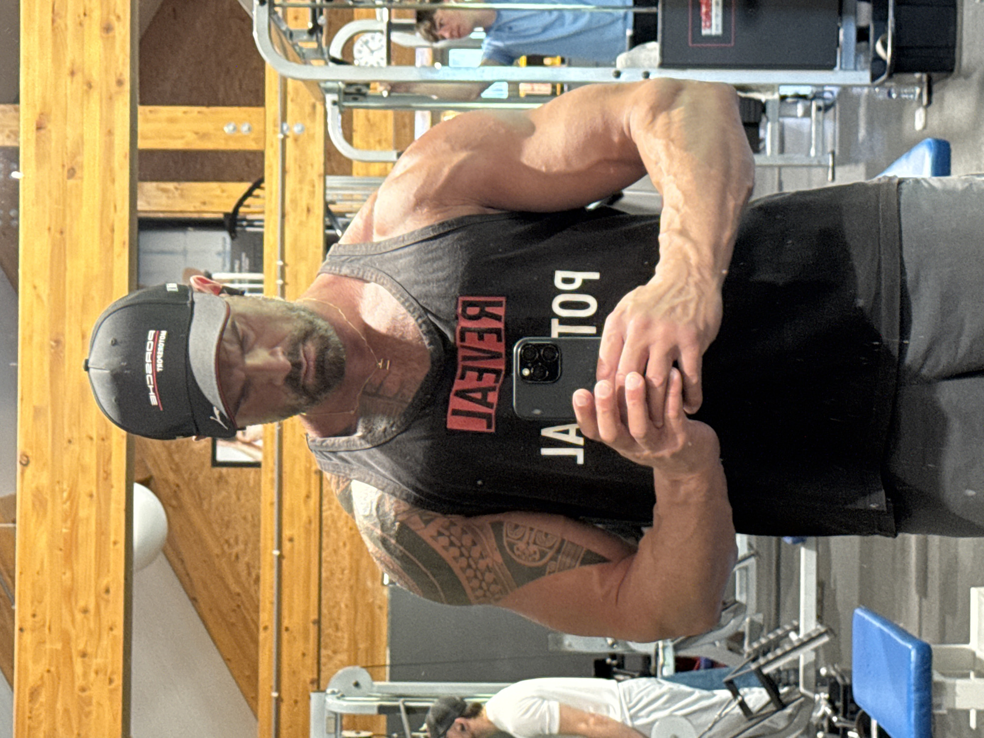 A man with a beard and a black cap is taking a selfie in what appears to be a gym. He is wearing a black tank top with the word "REVEAL" printed vertically on it, which is backwards due to the mirror reflection. He has tattoos on his arm and is holding a cell phone in his hand. The gym has standard equipment and other people working out. The location is unclear, and there is no information given about Jemelle, Belgium.
