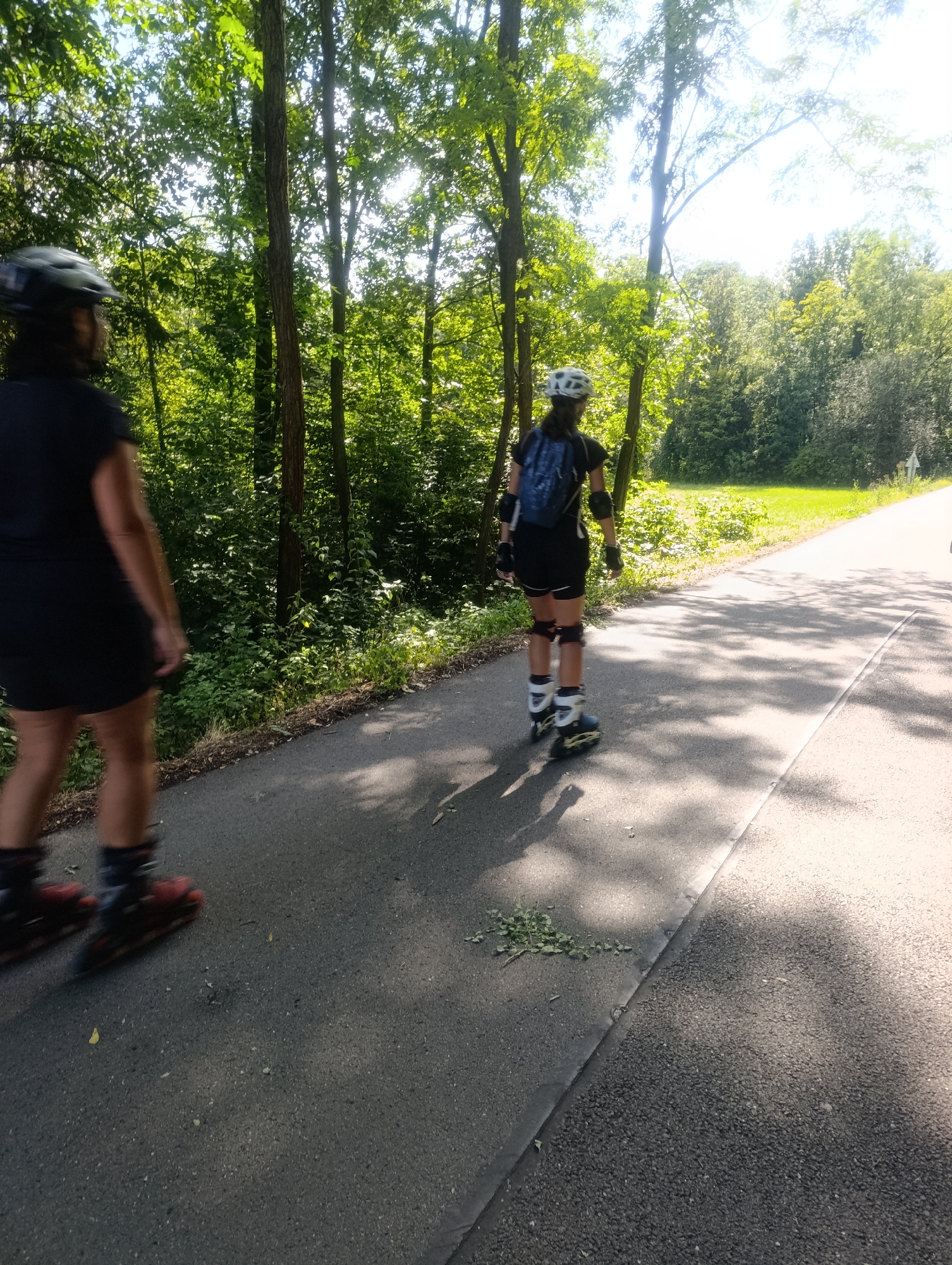 Two individuals are rollerblading on a paved path in a park-like setting. The person in the foreground is closer to the left edge of the frame and is wearing a black dress, helmet, and rollerblades. Their movement is blurred, indicating motion. The second person is further ahead, on the right side of the path, and is also wearing a helmet, a black t-shirt and shorts, knee pads, wrist guards, and rollerblades. They are carrying a blue backpack. The path is bordered by trees and lush green foliage, suggesting a wooded area. Sunlight filters through the leaves, casting shadows on the path. The scene appears to be outdoors on a sunny day. The specific location in Olomouc, Czechia, is not discernible from the image itself.