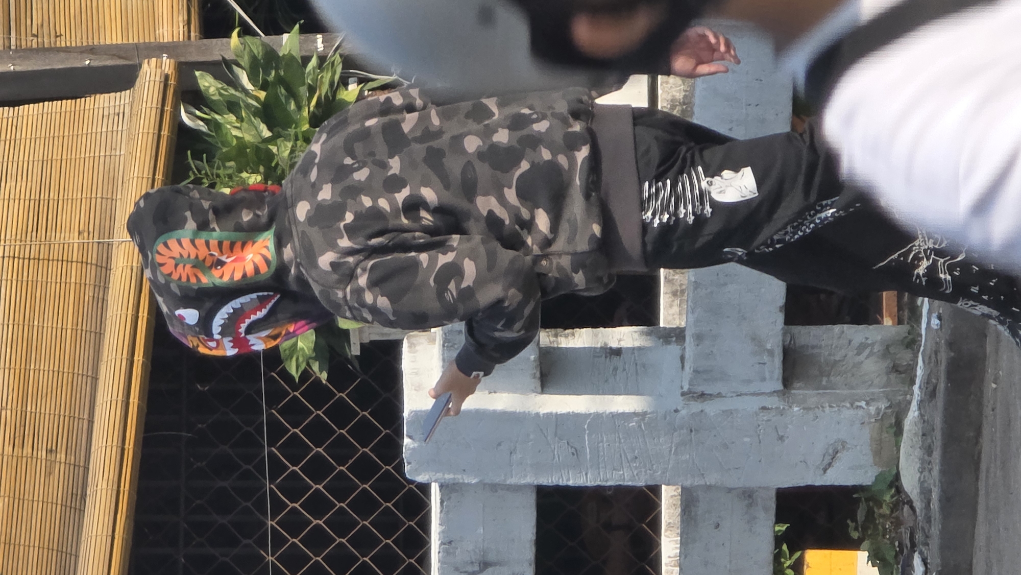 In Bắc Nha Trang, Vietnam, an individual wearing a camouflage hooded sweatshirt with a shark design on the hood and black pants with unique white patterns is seen holding a smartphone. The person stands near a fence and a concrete structure.