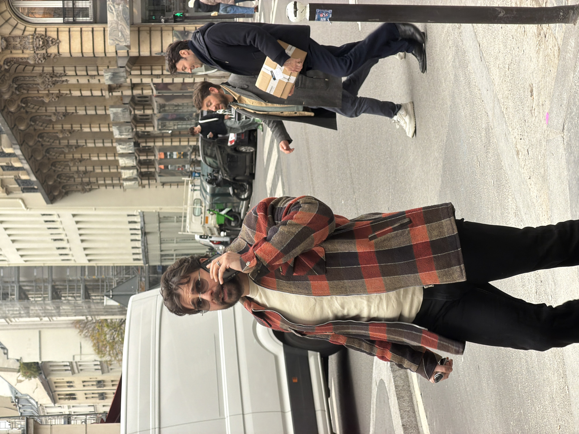 A street scene in Paris, France, shows a man in a plaid shirt and black pants talking on a cellphone, with a white van behind him. Two other men in the background are walking in the opposite direction; one carries a small package. The background includes buildings with ornate architectural details, parked vehicles, and a street with pedestrian markings.
